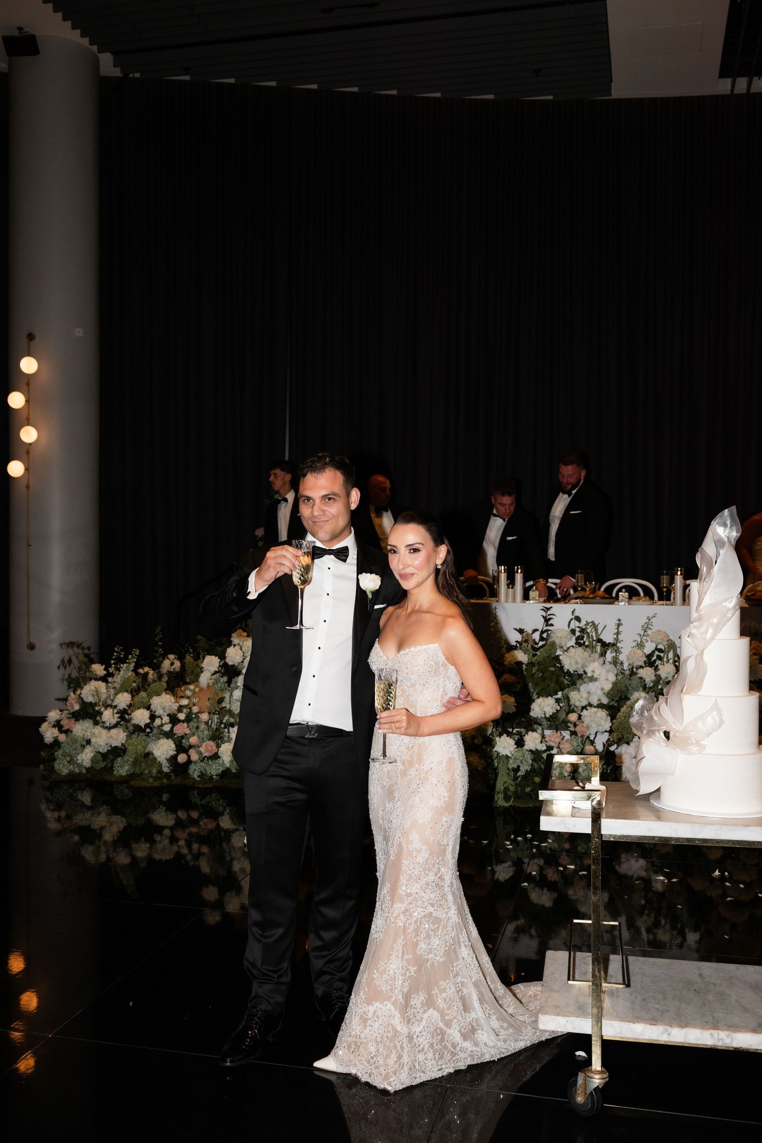 Bride and groom toasting with champagne glasses near wedding cake and floral arrangements at elegant reception.