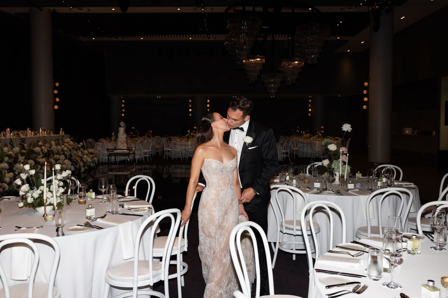 Bride and groom share a kiss in an elegantly decorated wedding reception venue with white floral centerpieces.