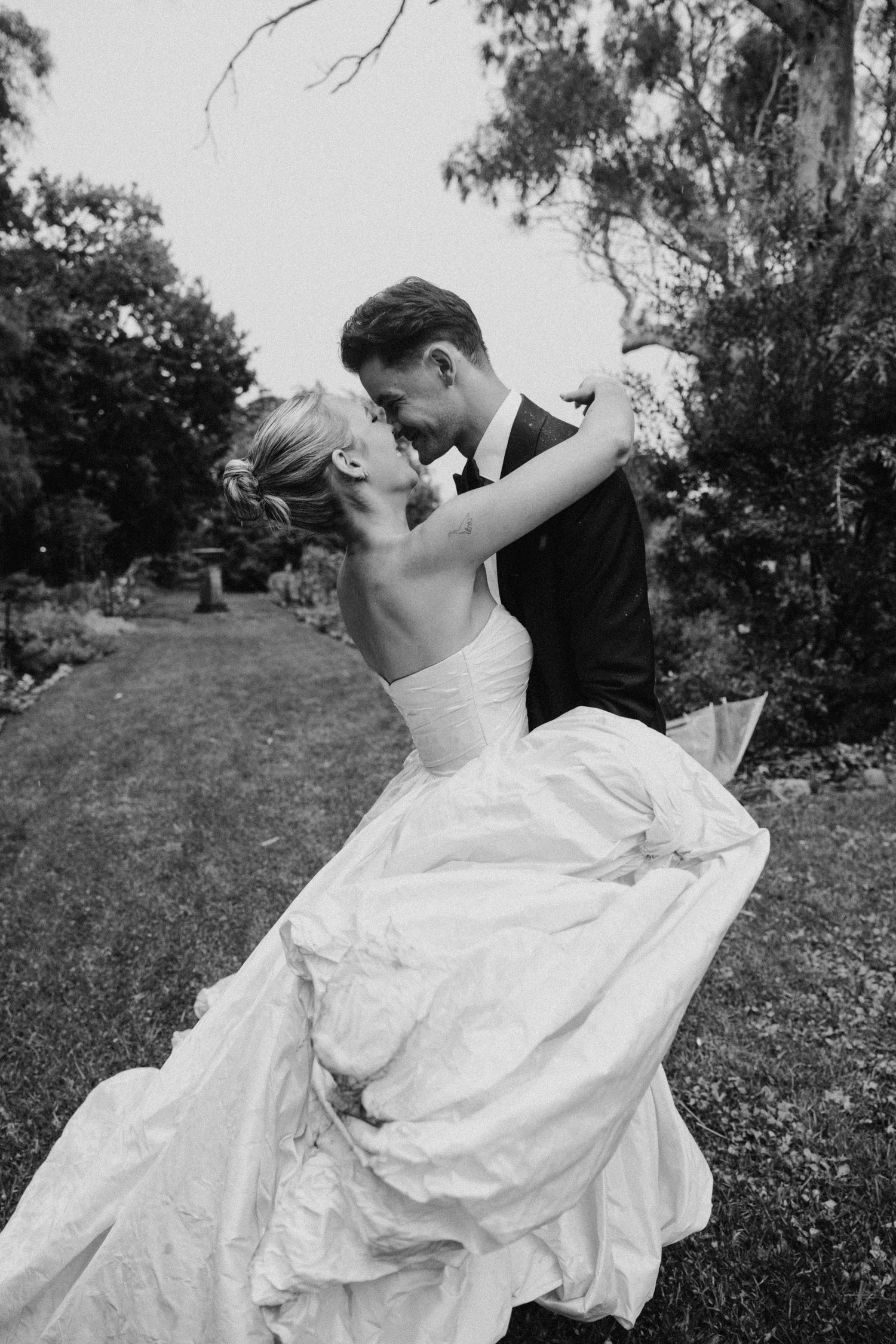 Black and white photo of a bride and groom sharing a romantic dip kiss in a lush garden on their wedding day.