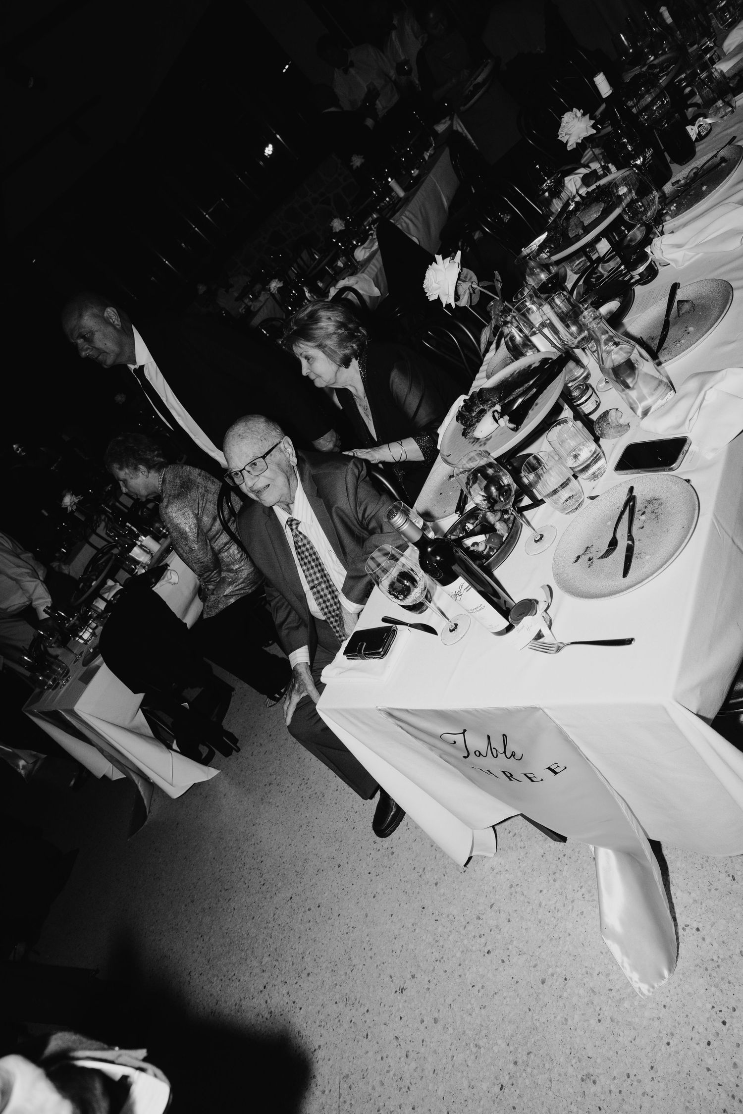 Black and white photo of elegantly dressed guests mingling at a formal dinner gala event with decorated tables.