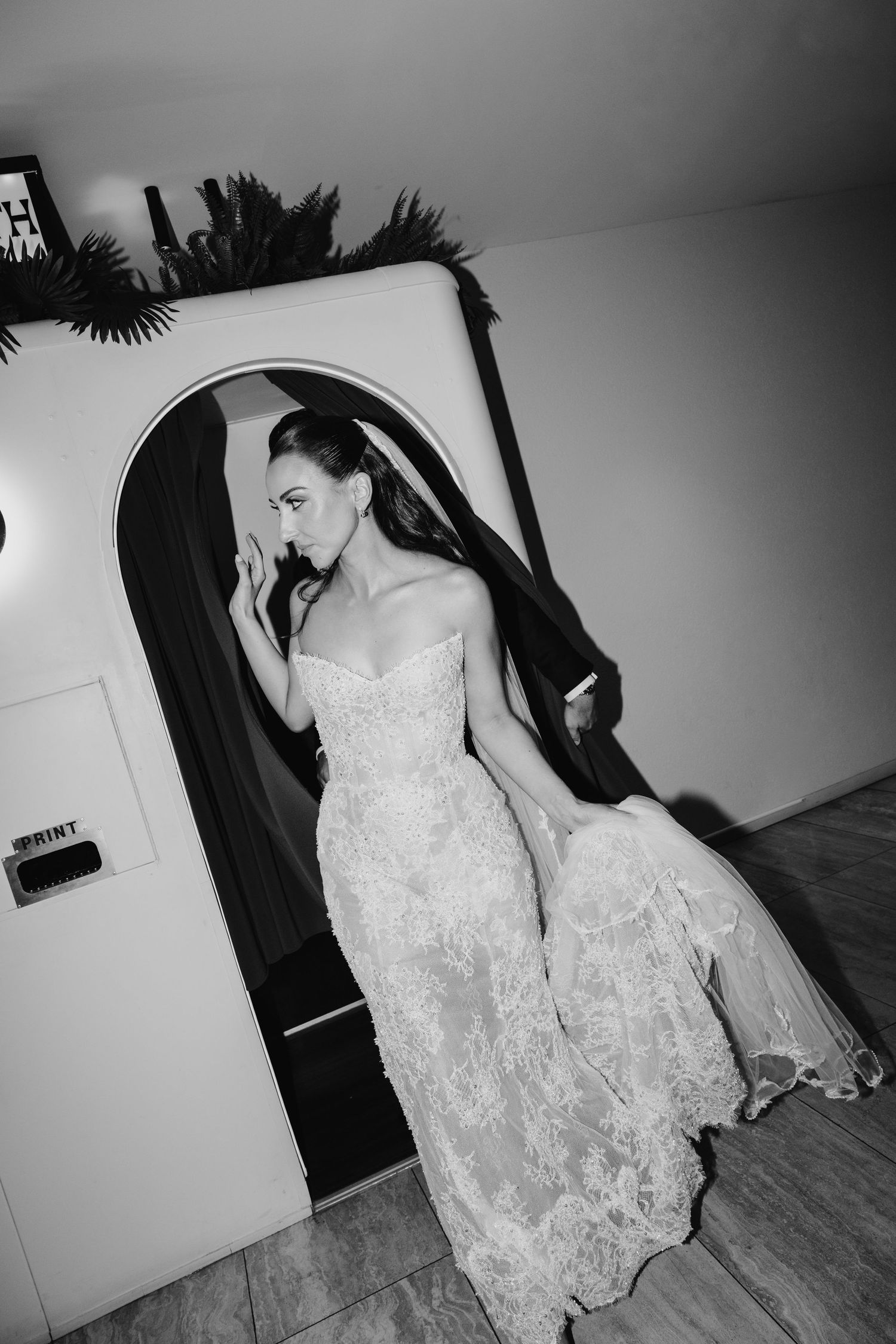 Bride in strapless lace wedding gown poses elegantly in black and white photo near an arched doorway.