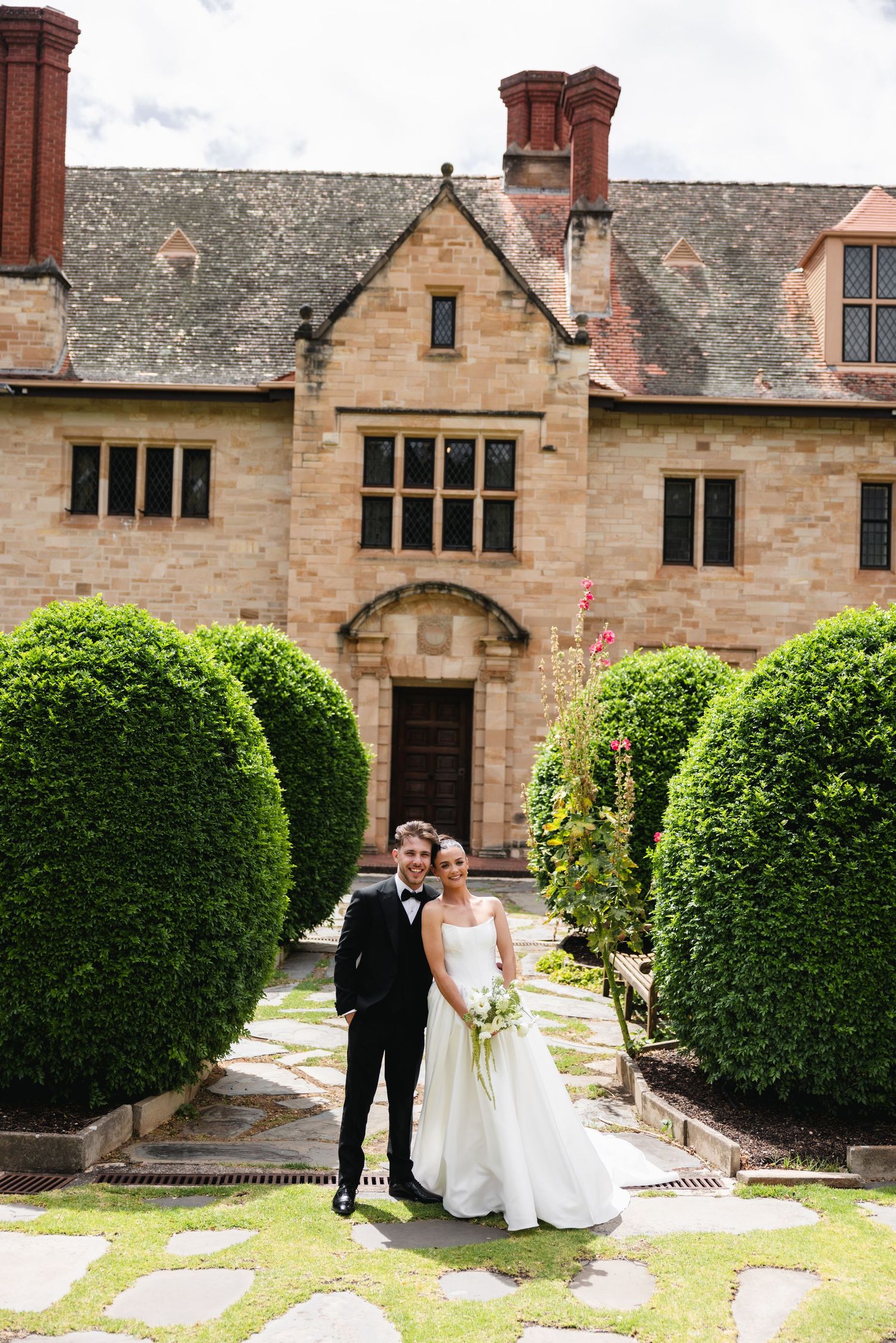 Bride and groom pose together in formal garden with manicured hedges before a grand stone manor house.