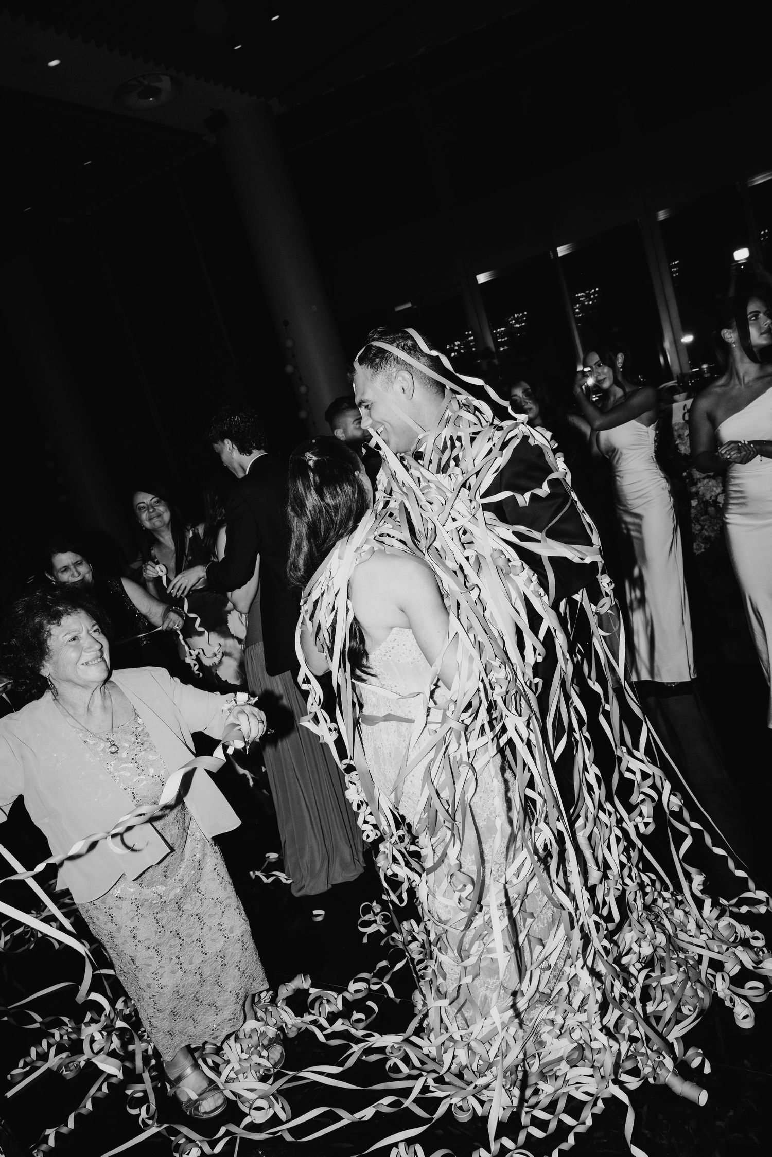 Groom covered in streamers dancing at wedding reception in black and white photo with guests watching.