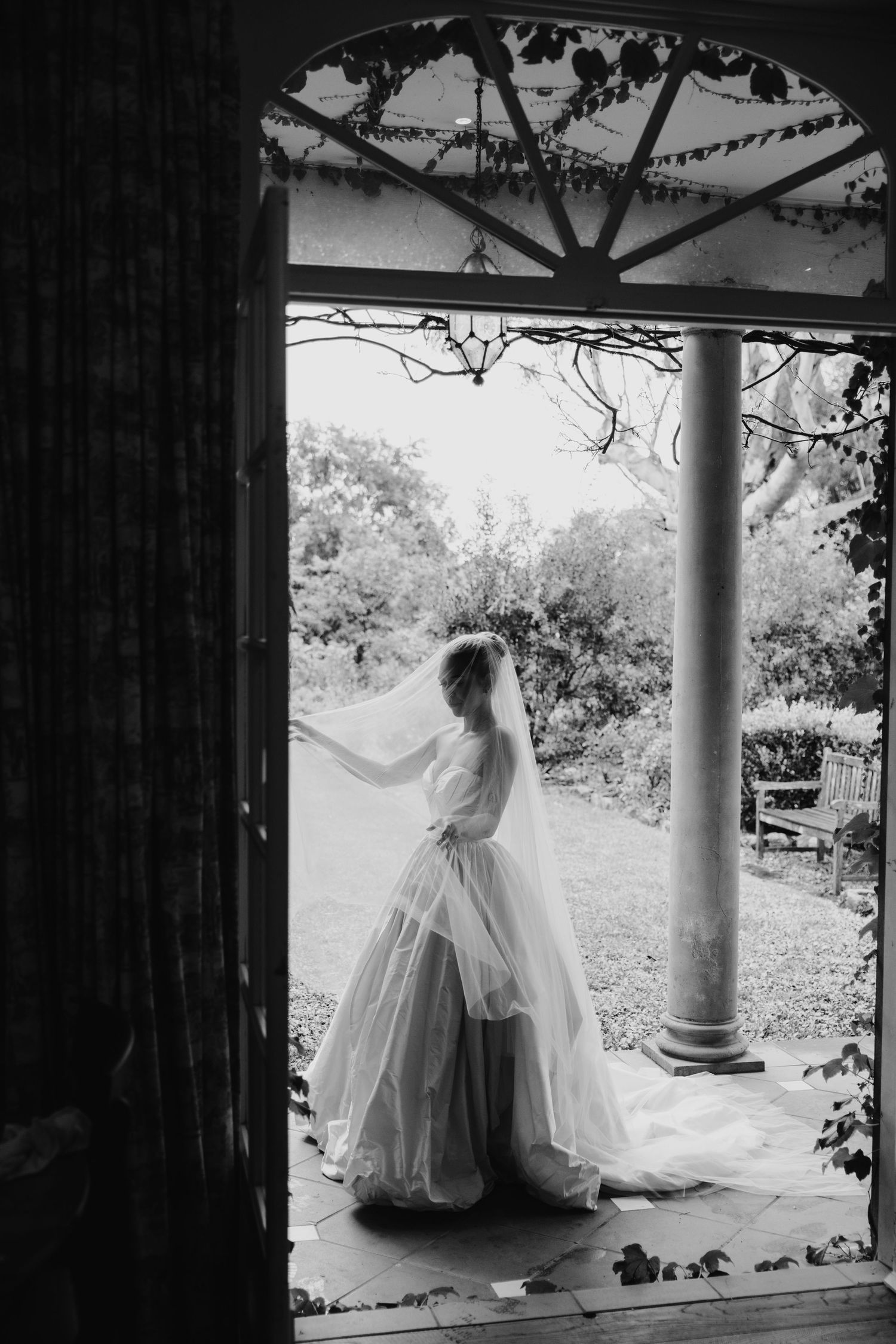 Elegant bride in tulle wedding gown stands in doorway of vintage venue in black and white photo.
