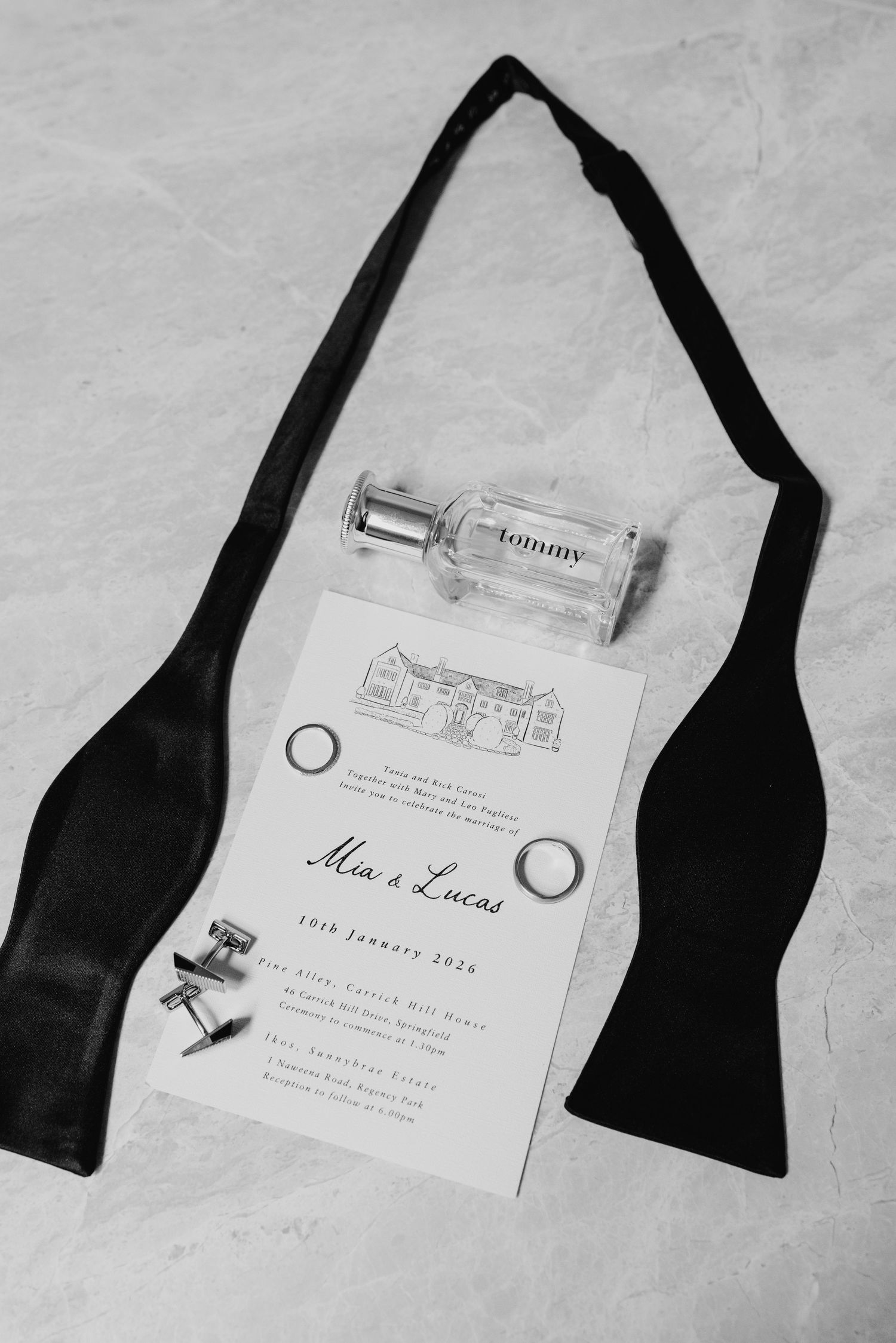 Black and white flat lay of groom wedding accessories including bow tie, Tommy cologne, cufflinks, rings, and elegant invitation.