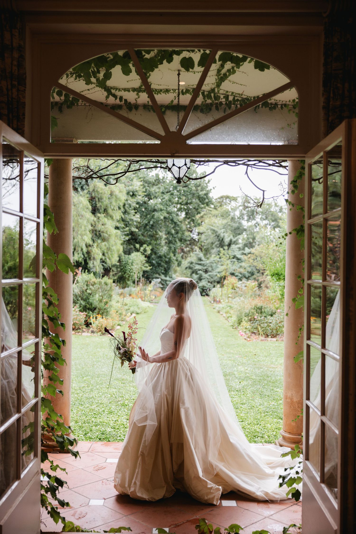 Bride in a flowing white gown and veil standing in a vine-covered doorway overlooking a lush garden.