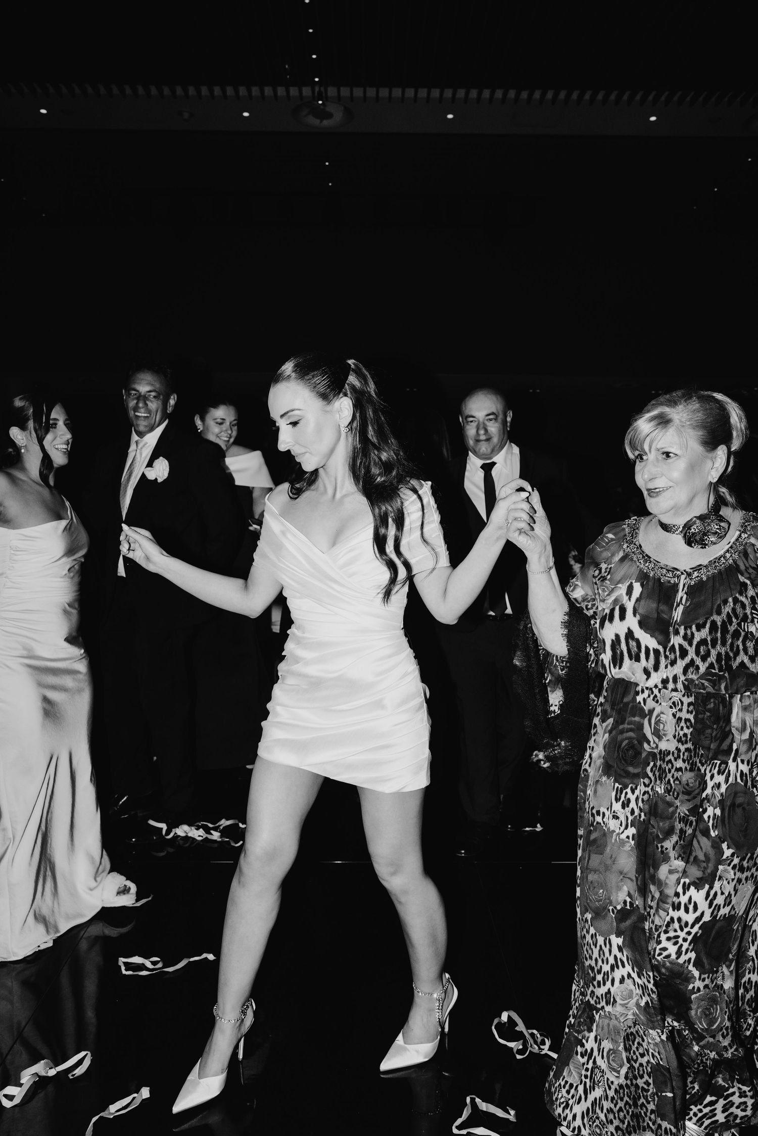 Bride in short white dress dancing with elderly guest at wedding reception in black and white photo.