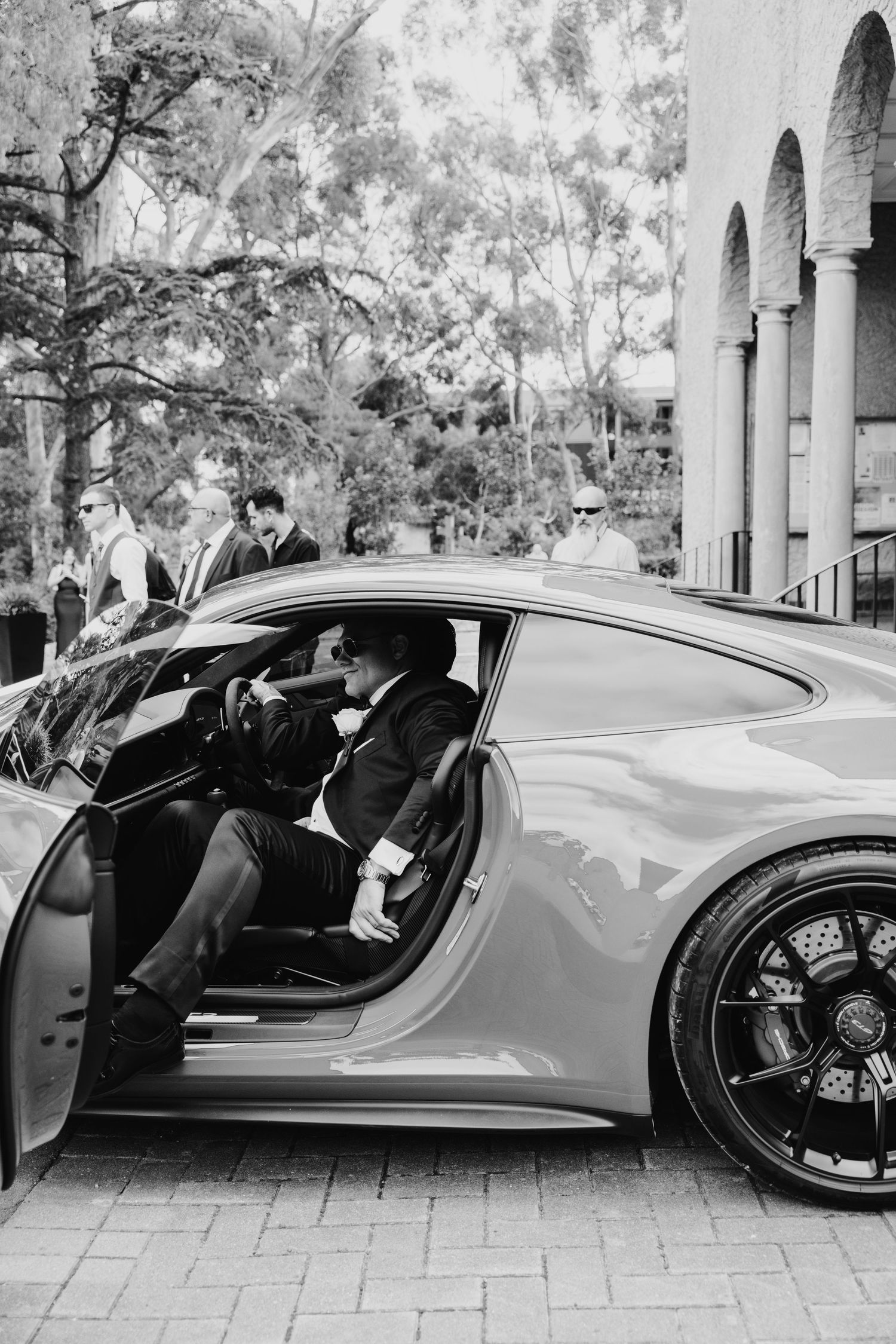 Groom in black tuxedo sitting in luxury sports car on wedding day.