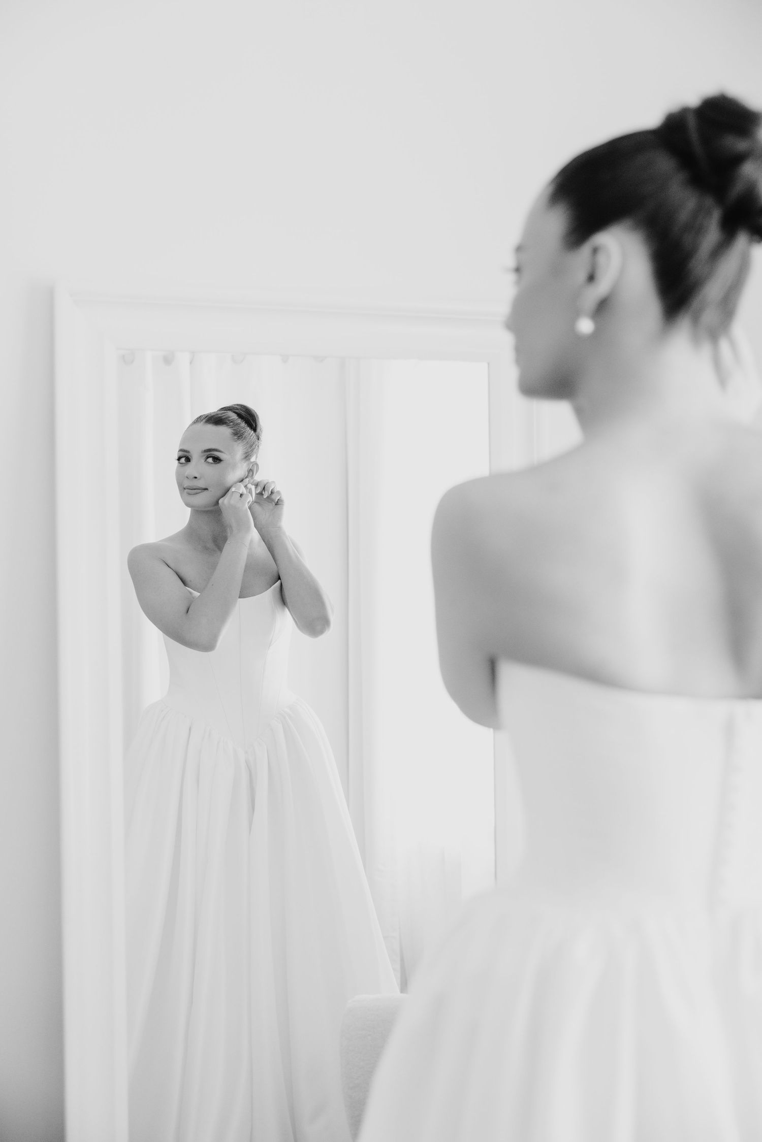 Bride in strapless white wedding gown adjusting earring while looking at her reflection in a mirror.
