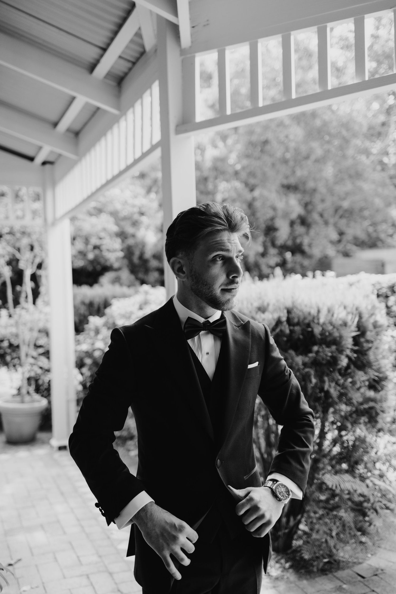 Dapper groom in black tuxedo and bow tie poses confidently on a porch surrounded by lush greenery.