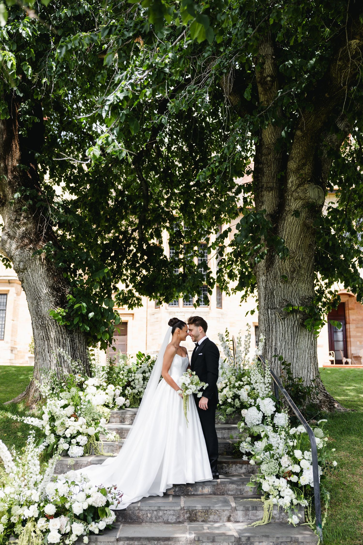Bride and groom share romantic moment on flower-lined steps beneath lush trees at elegant outdoor wedding venue.