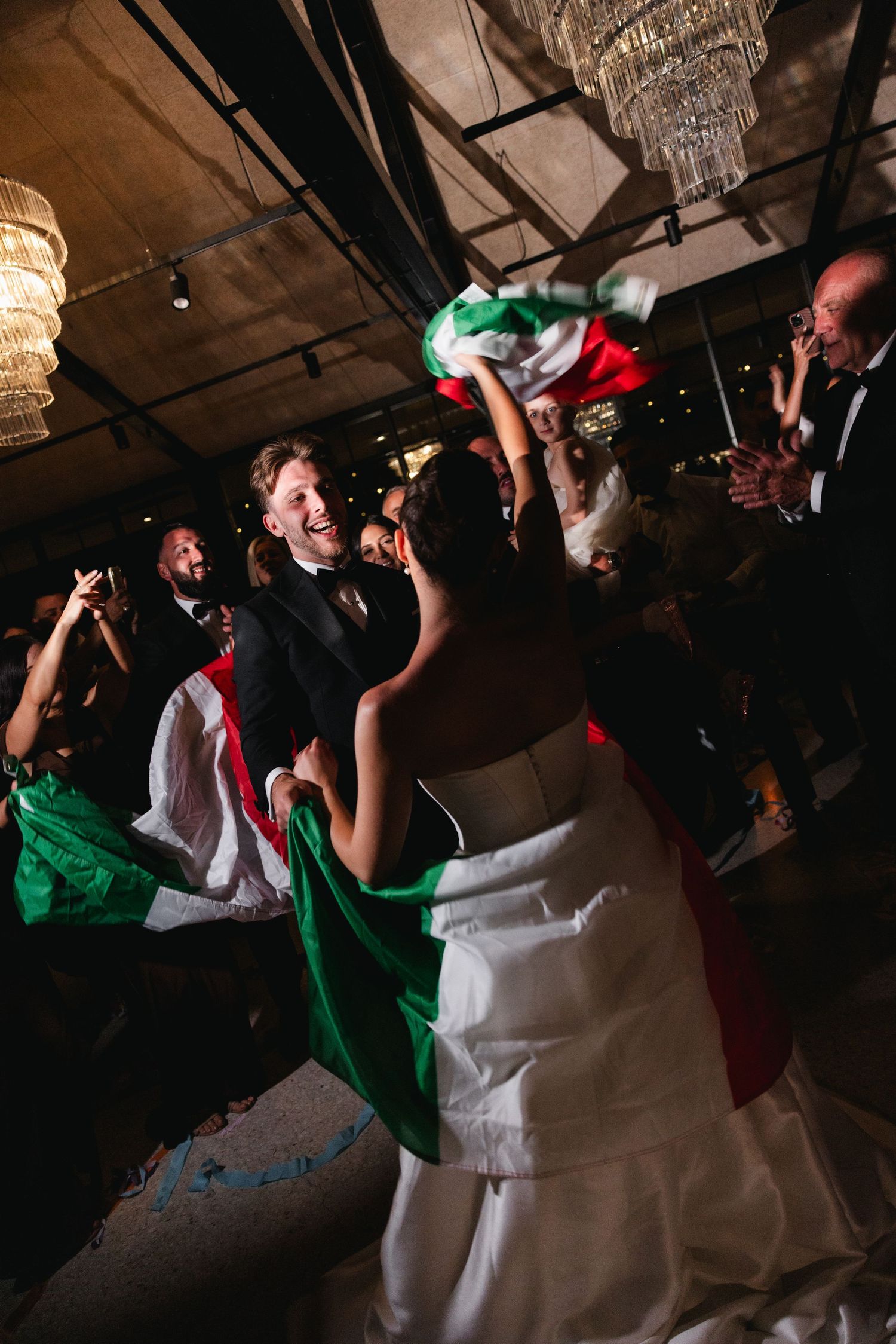 Joyful couple celebrates wedding reception dancing with Italian flags surrounded by cheering guests under chandeliers.