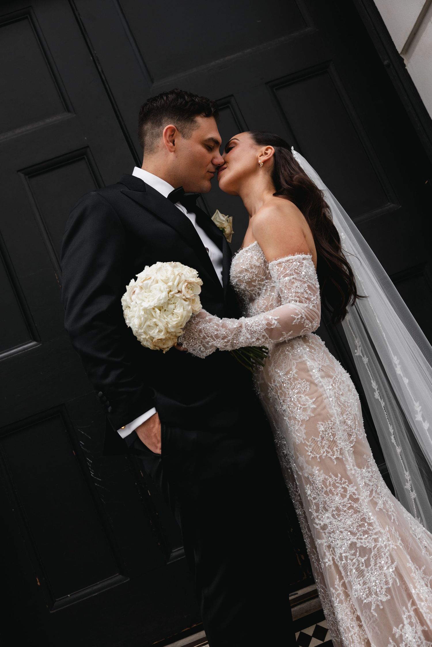 Bride and groom share a romantic kiss in front of black doors, bride wearing lace gown holding white bouquet.