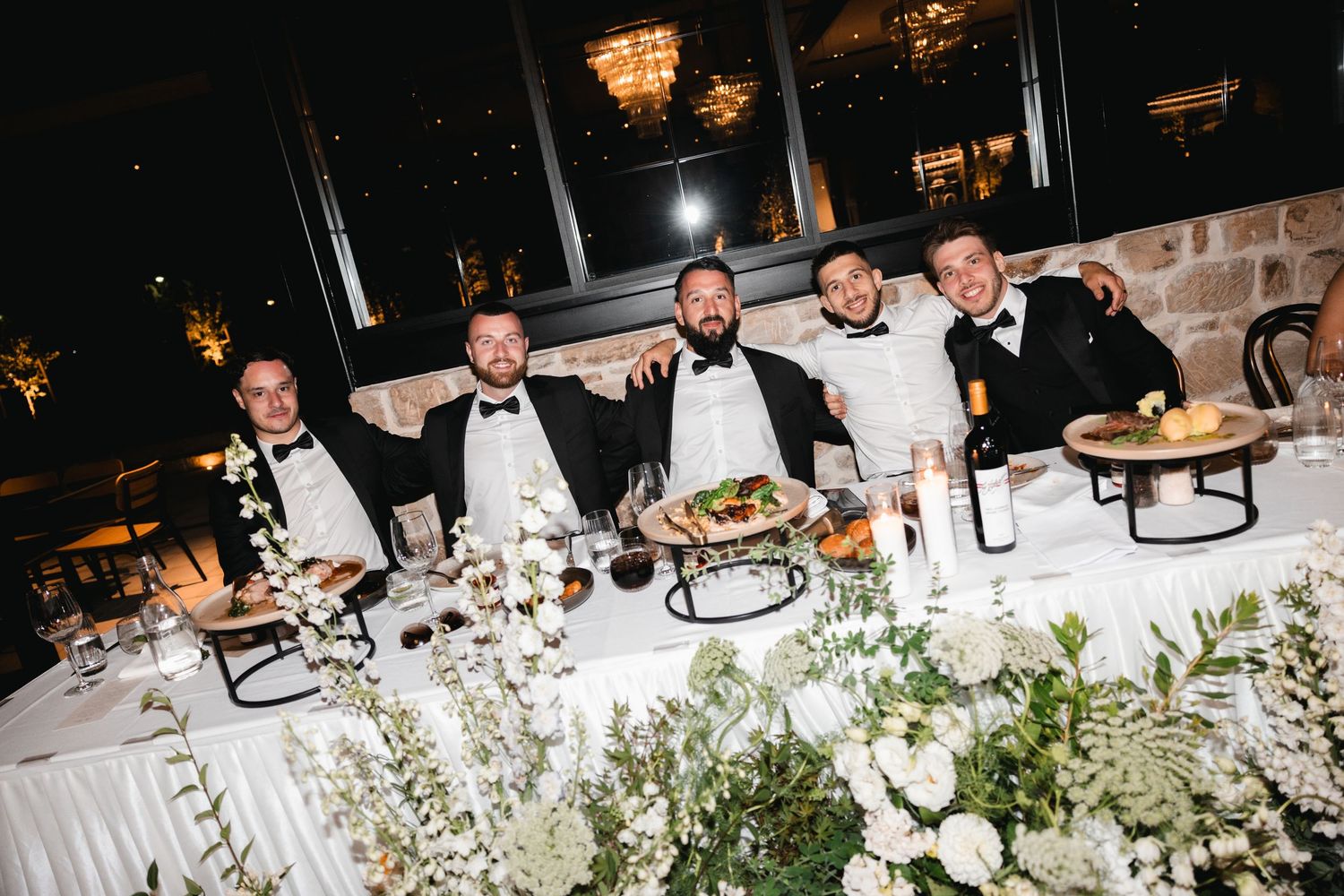 Five men in tuxedos smile at a elegantly decorated wedding reception table with white floral arrangements.