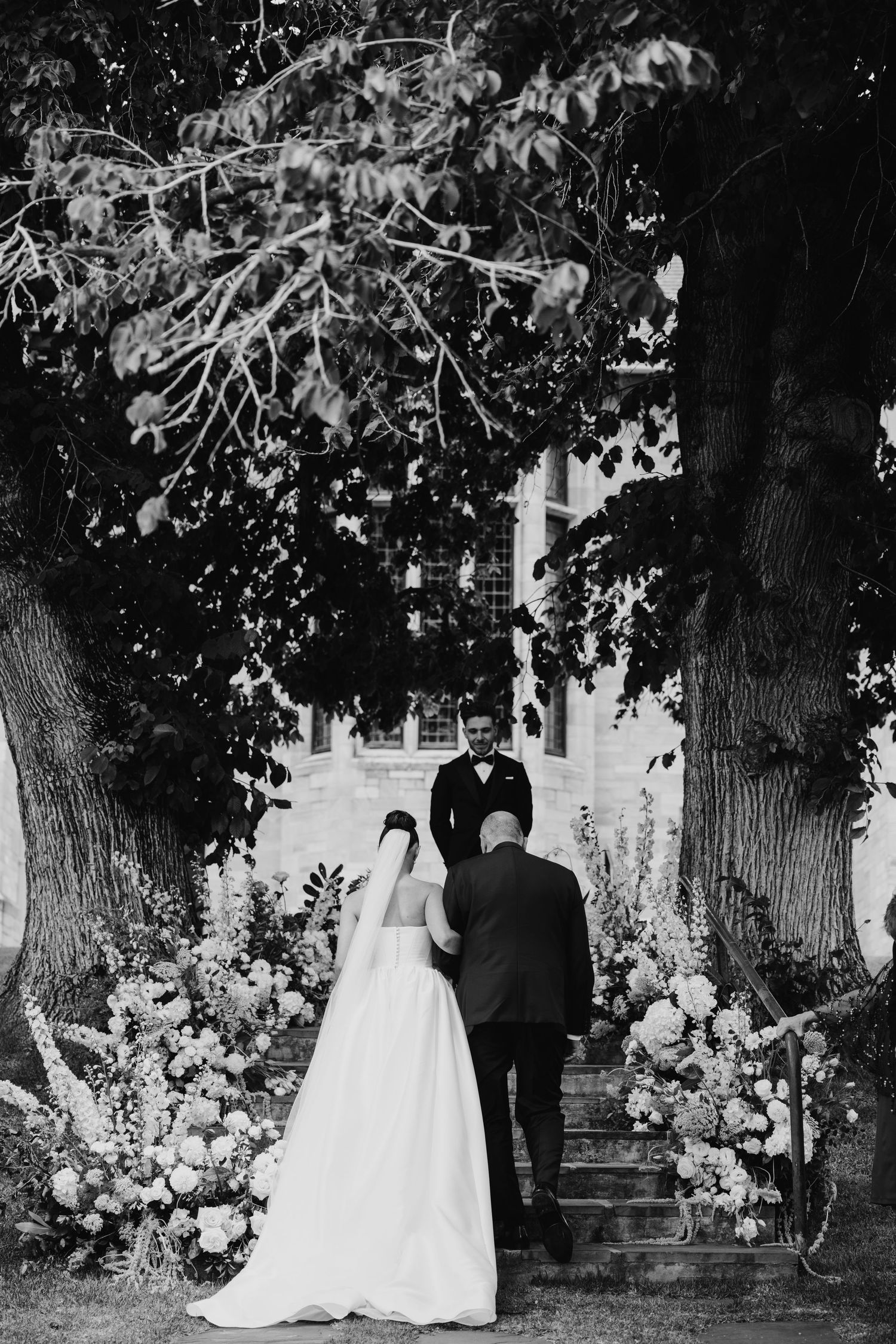 Black and white wedding ceremony photo of bride and groom standing beneath large trees surrounded by elegant floral arrangements.