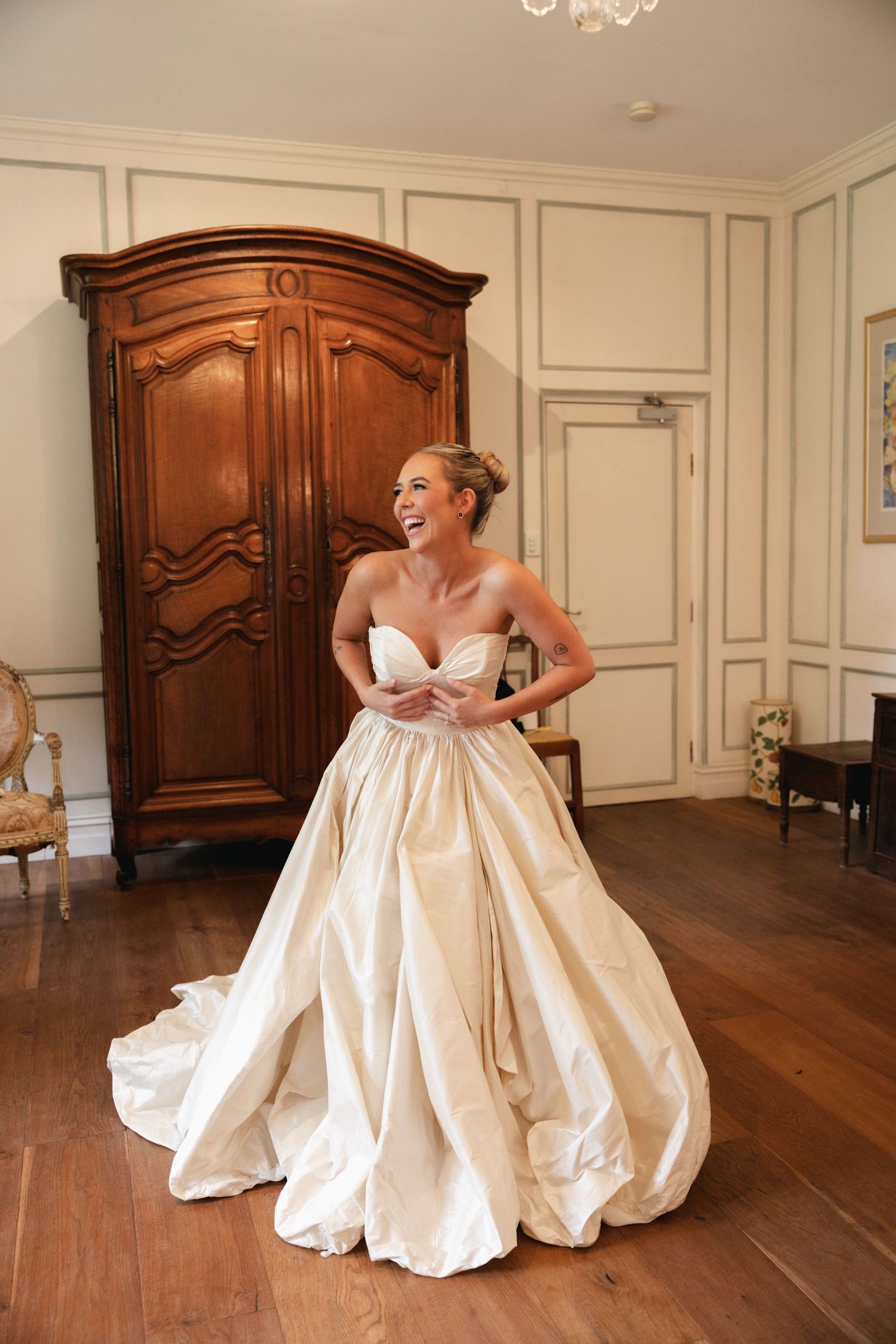 Smiling bride in strapless champagne ballgown wedding dress standing in elegant room with wooden armoire.