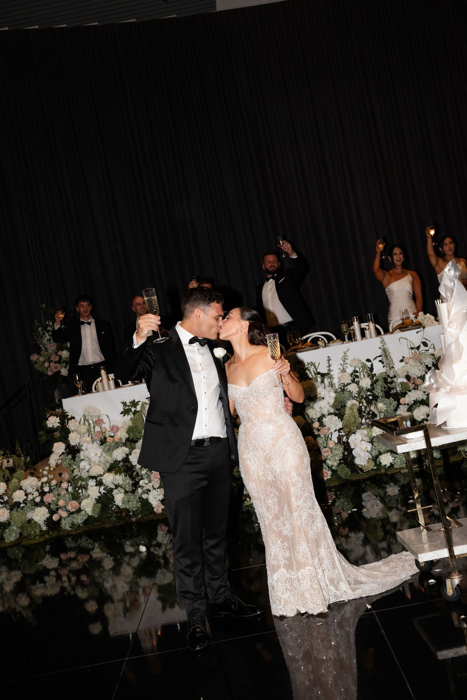 Bride and groom share a champagne toast kiss at elegant wedding reception with floral arrangements and wedding party behind them.