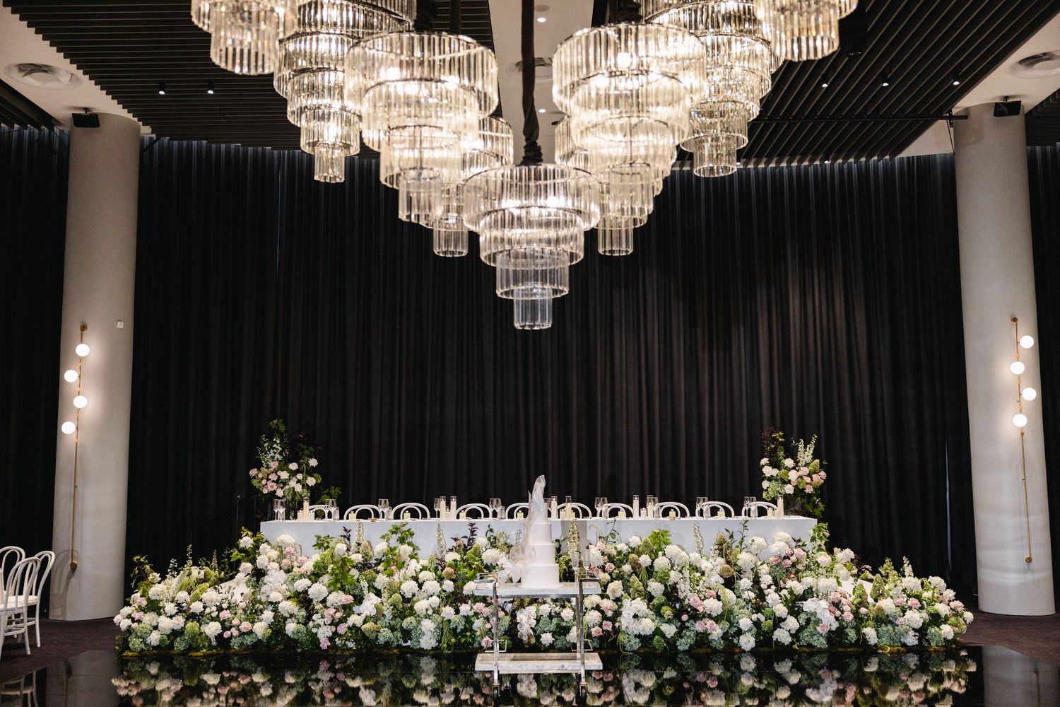 Elegant wedding reception with tiered crystal chandeliers above a floral head table adorned with white blooms and greenery.