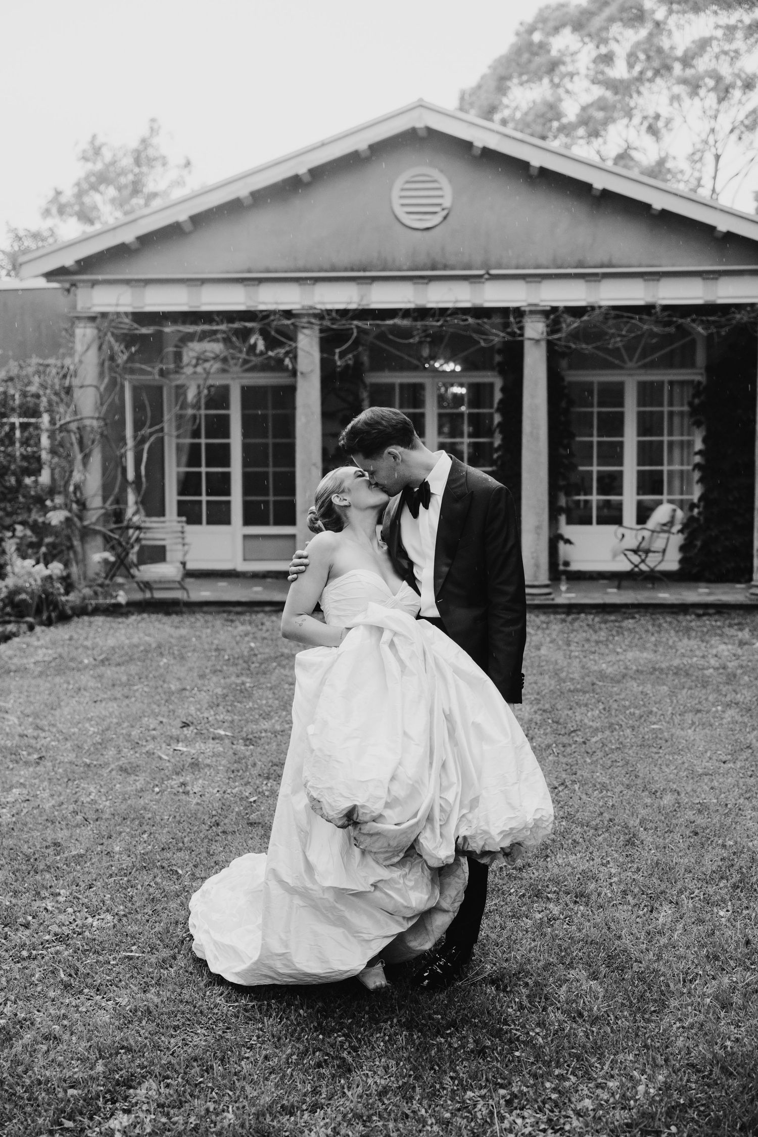 Black and white photo of a bride and groom kissing on a lawn in front of an elegant country house.