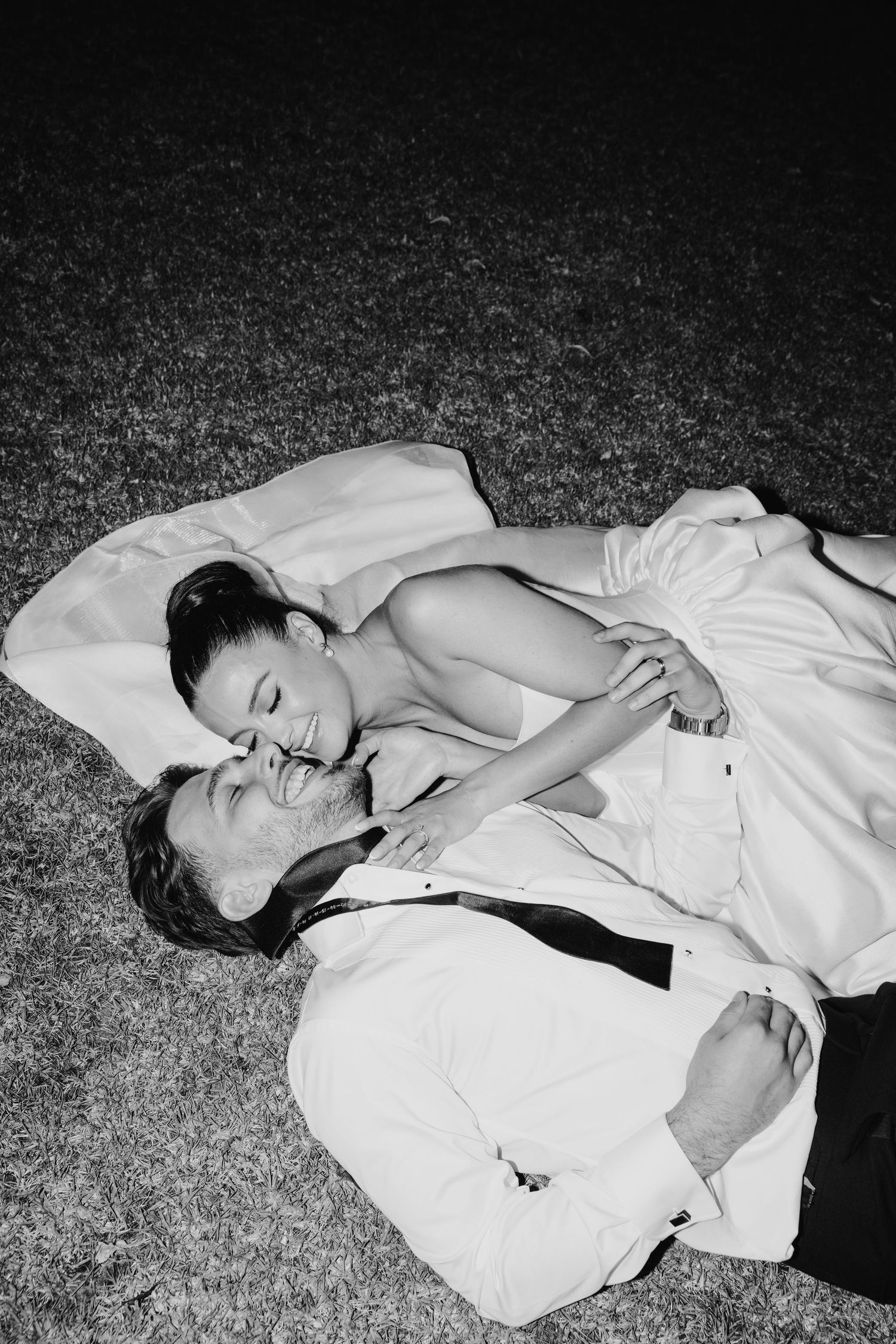 Black and white photo of a happy couple in wedding attire lying on grass, sharing a romantic moment.