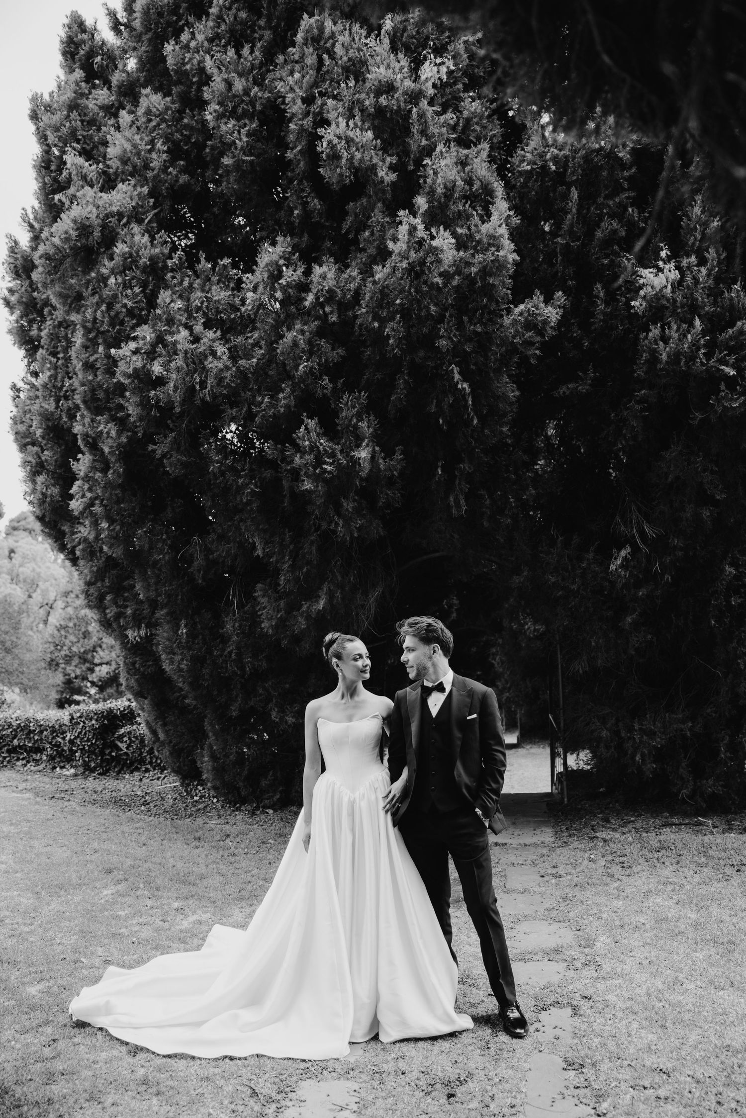 Black and white wedding photo of bride in flowing gown and groom in tuxedo standing before a large evergreen tree.
