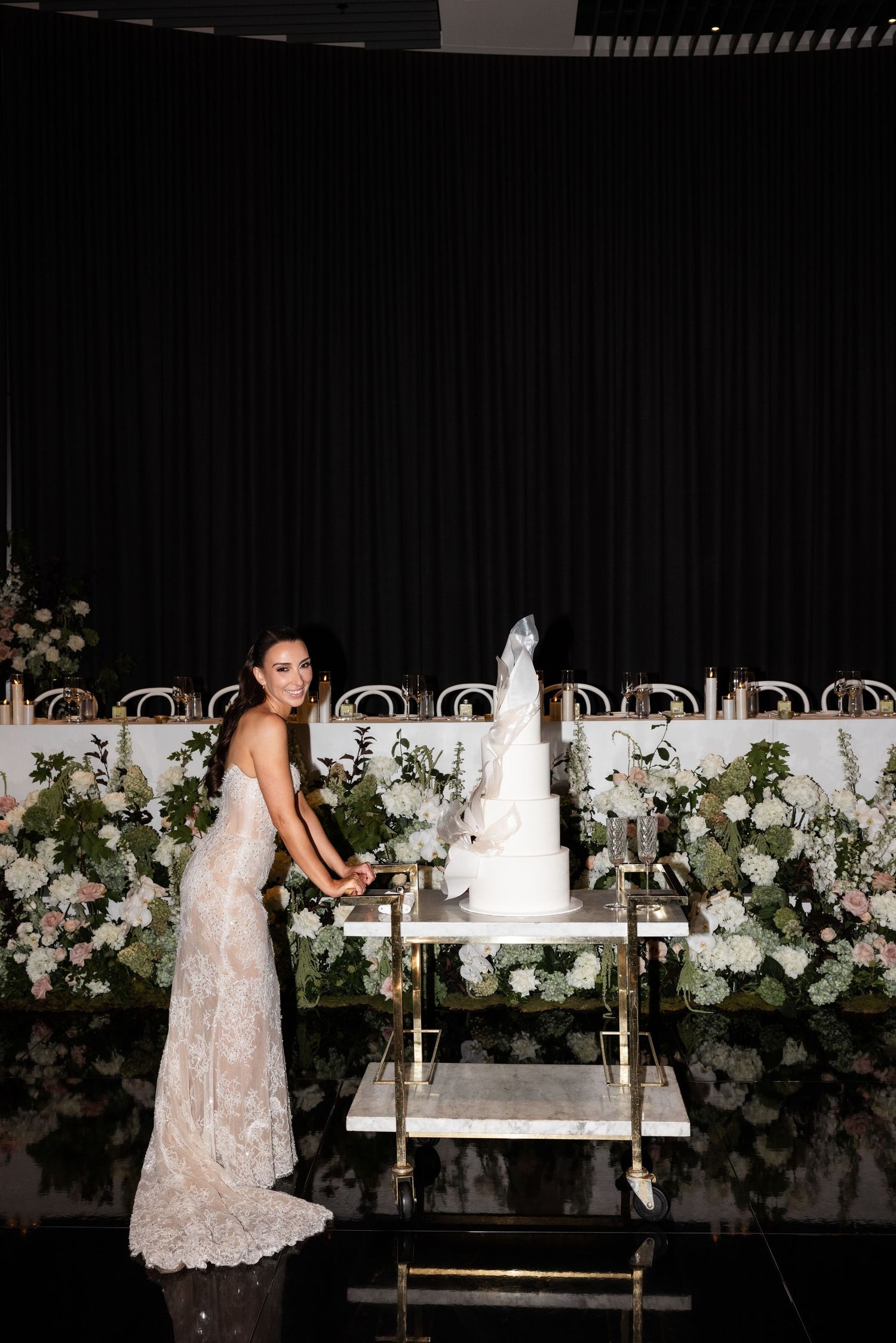 Bride in lace gown cutting elegant white multi-tiered wedding cake at floral-adorned reception.
