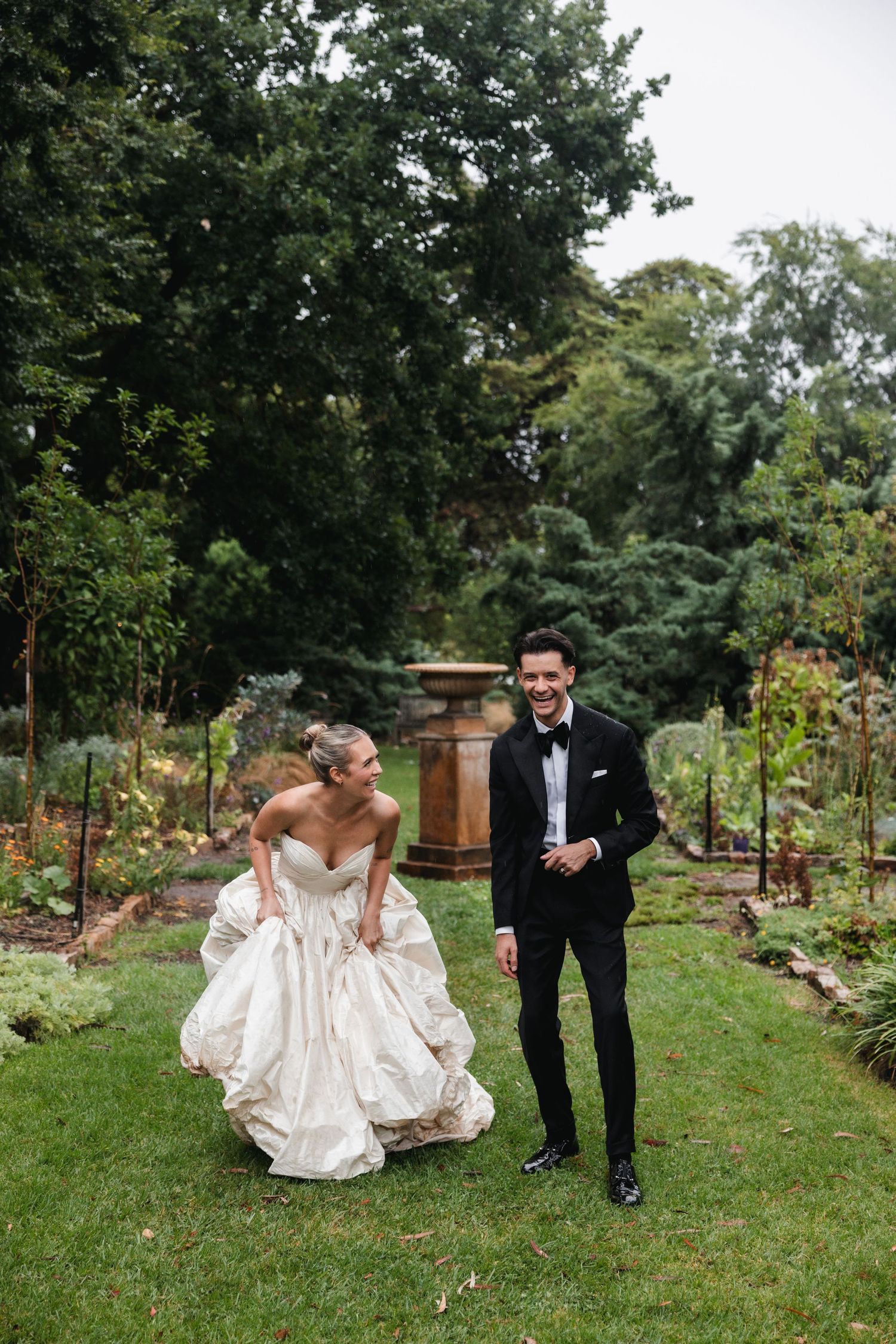 Bride in strapless ball gown and groom in black tuxedo laughing together in lush garden on wedding day.