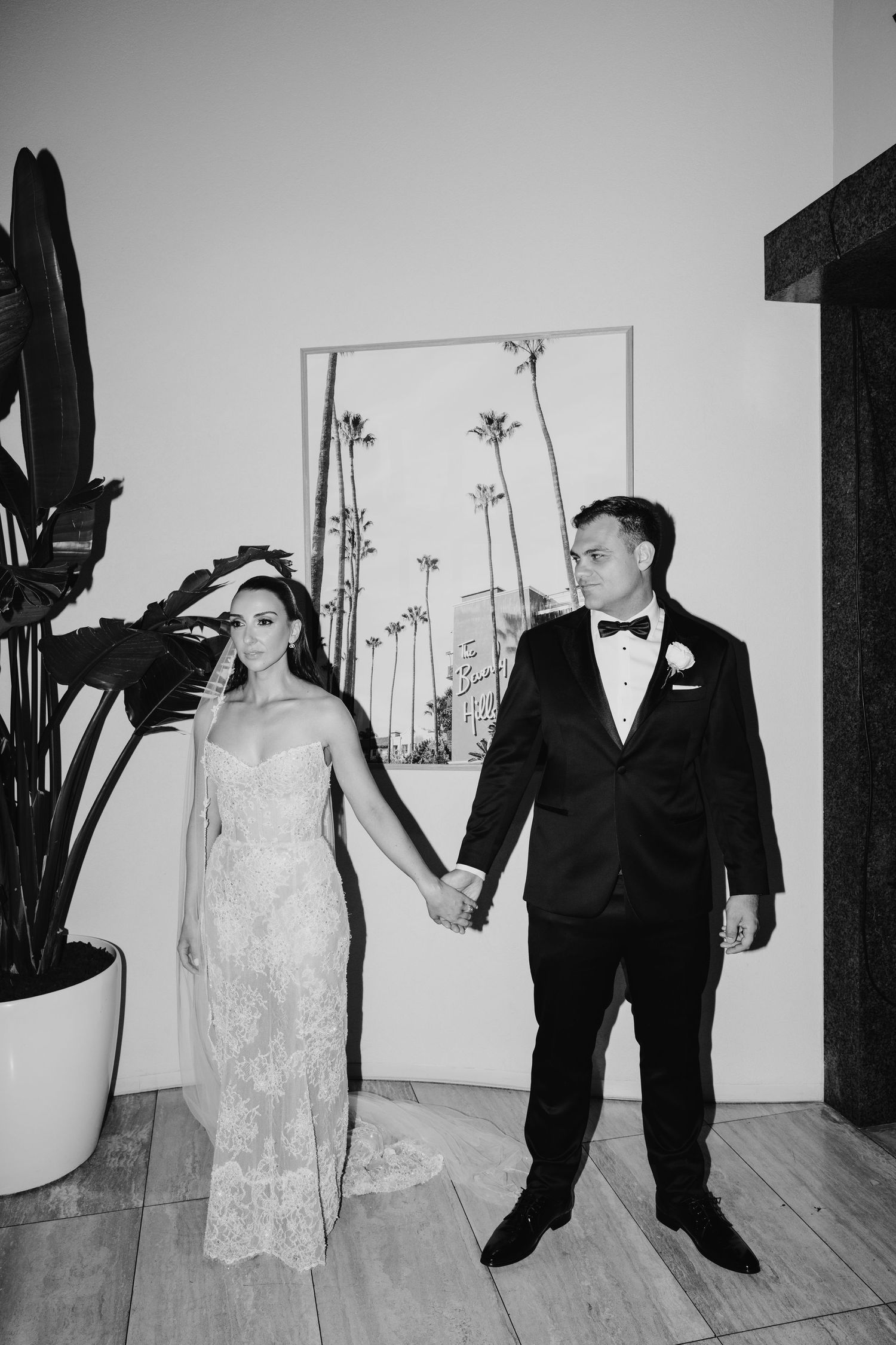 Black and white photo of bride and groom holding hands in elegant wedding attire with palm tree artwork behind them.