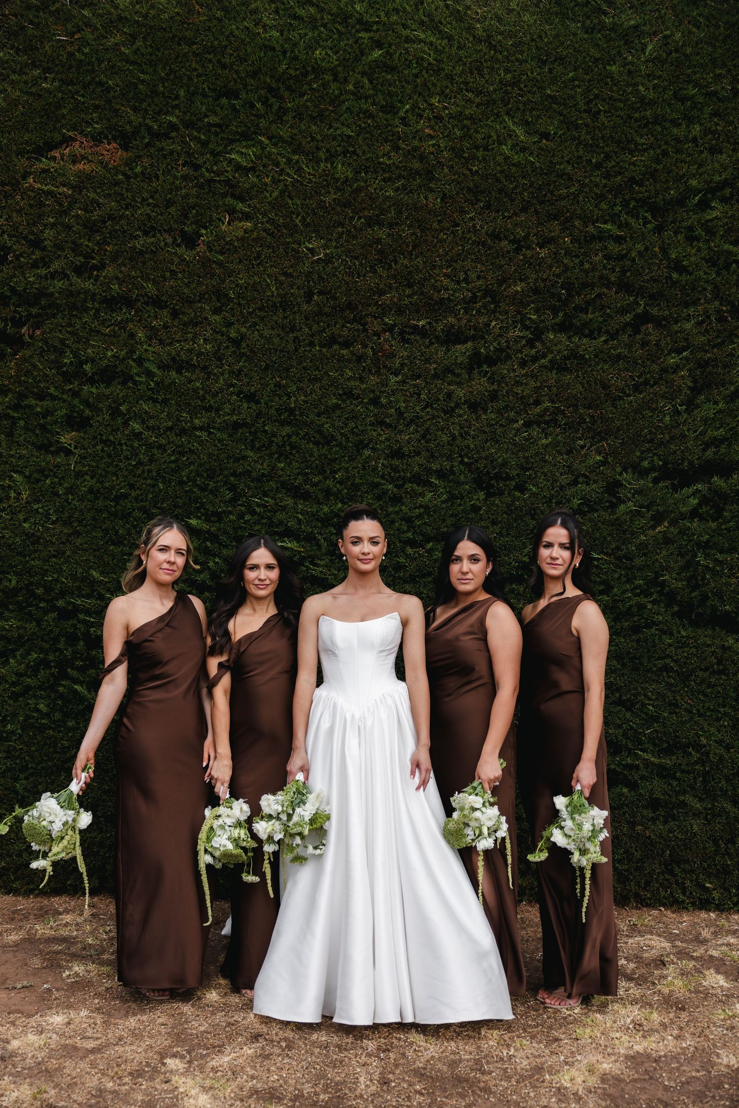 Bride in white gown poses with four bridesmaids in brown satin dresses holding white bouquets.