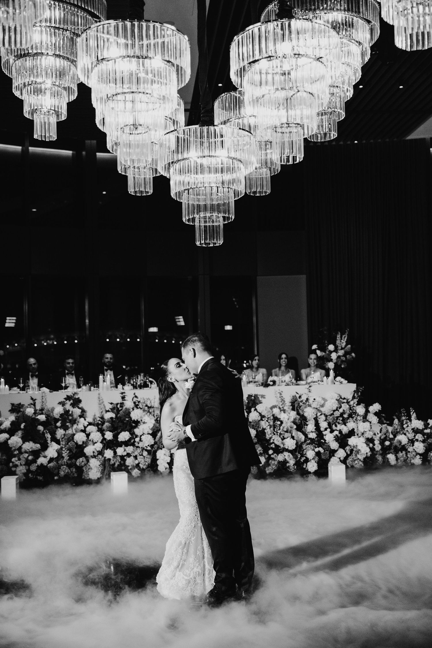 Black and white photo of couple sharing first dance under crystal chandeliers with dry ice fog.