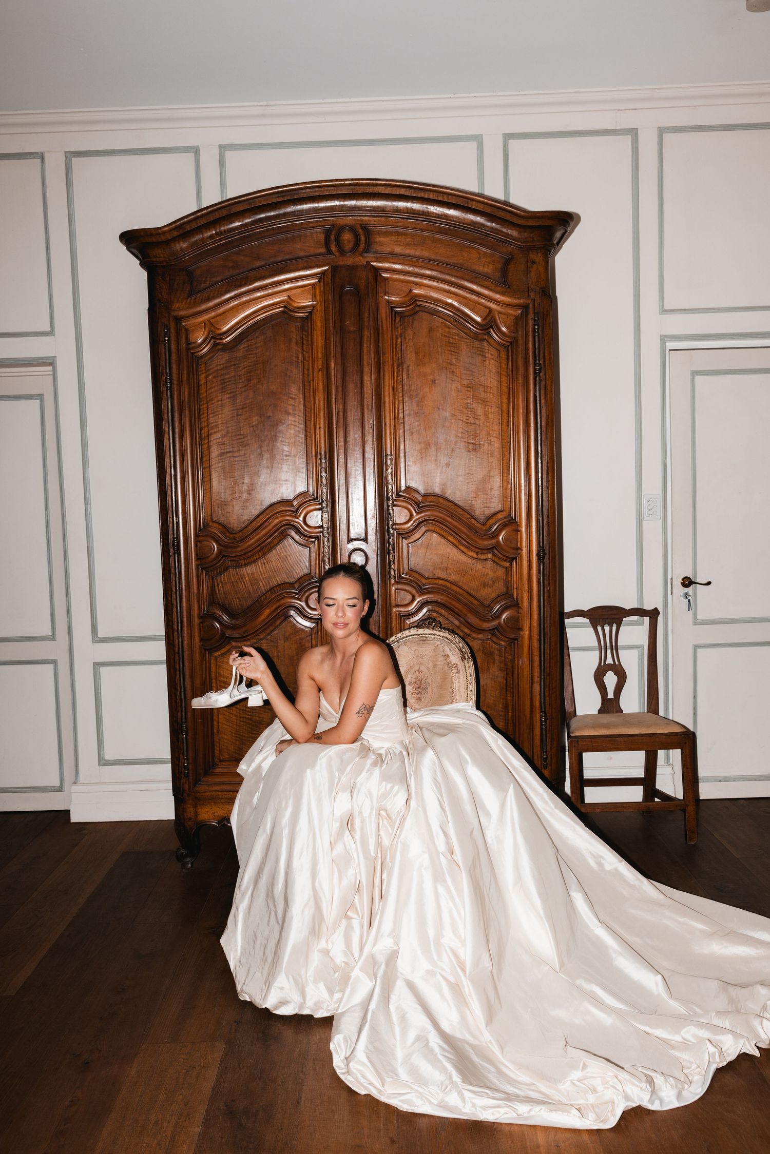 Bride in elegant satin ball gown sits before ornate wooden armoire, holding wedding shoe in classic bridal suite.