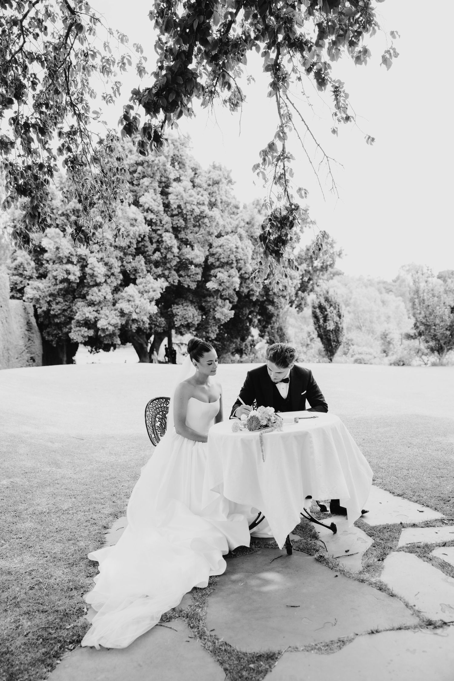 Black and white photo of bride and groom signing wedding documents at outdoor table beneath large trees.