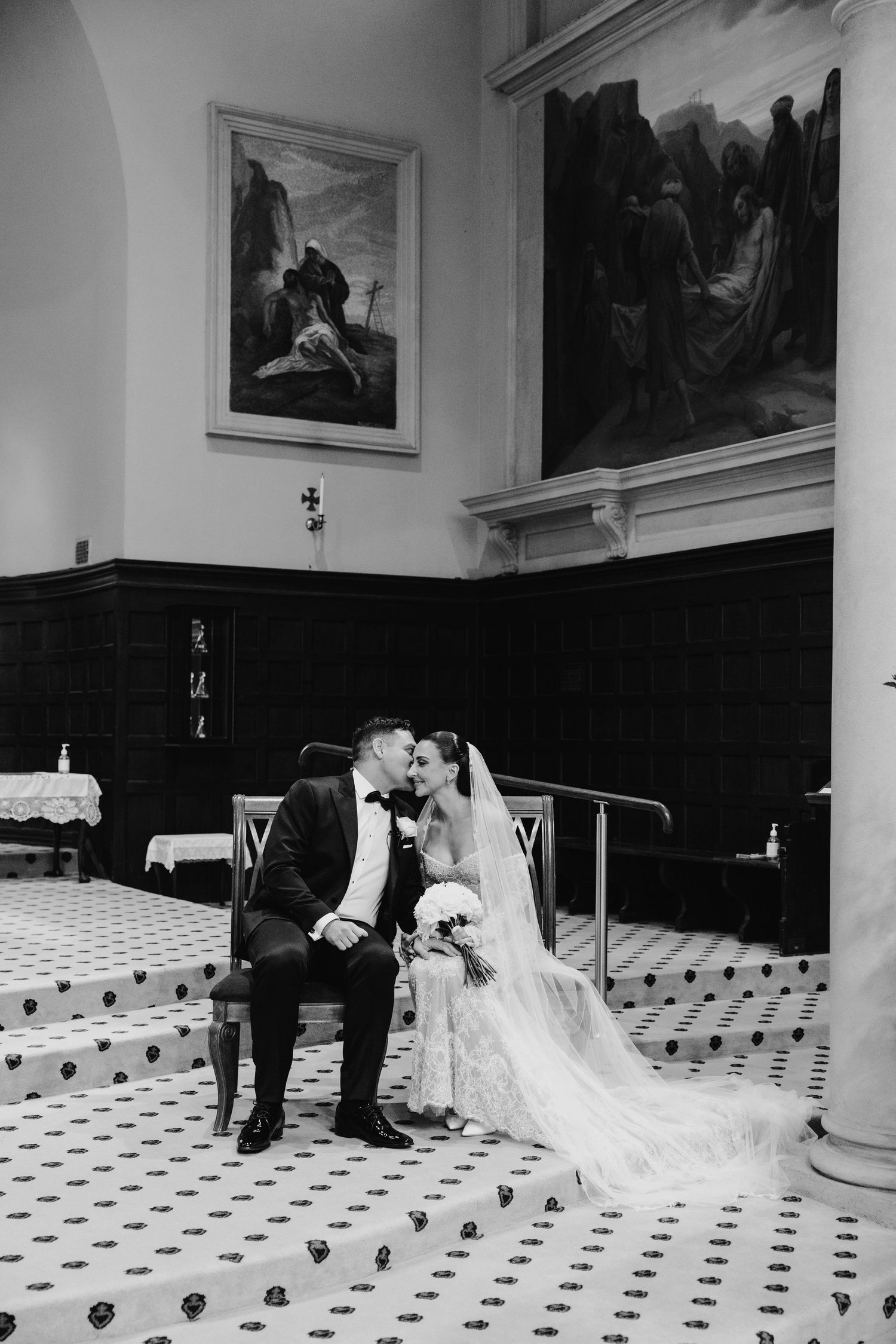 Black and white photo of a bride and groom sharing a kiss inside an elegant church with ornate paintings.