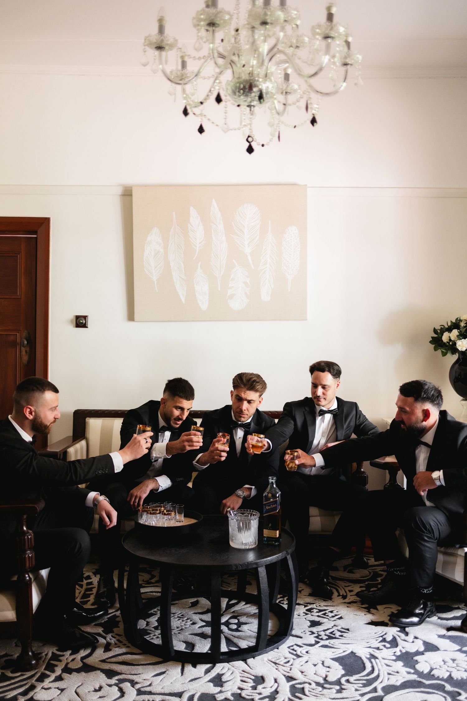 Five groomsmen in black tuxedos toasting with whiskey glasses around a table with a bottle of bourbon.