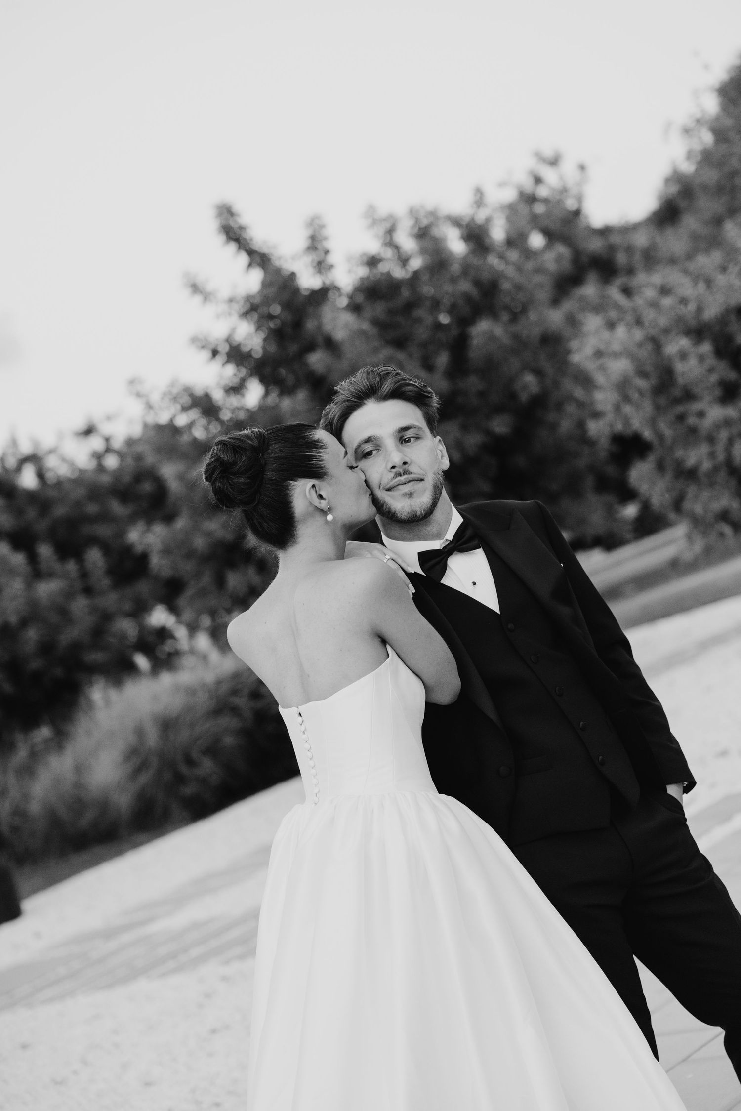 Black and white wedding photo of groom in tuxedo being kissed on cheek by bride in strapless gown outdoors.