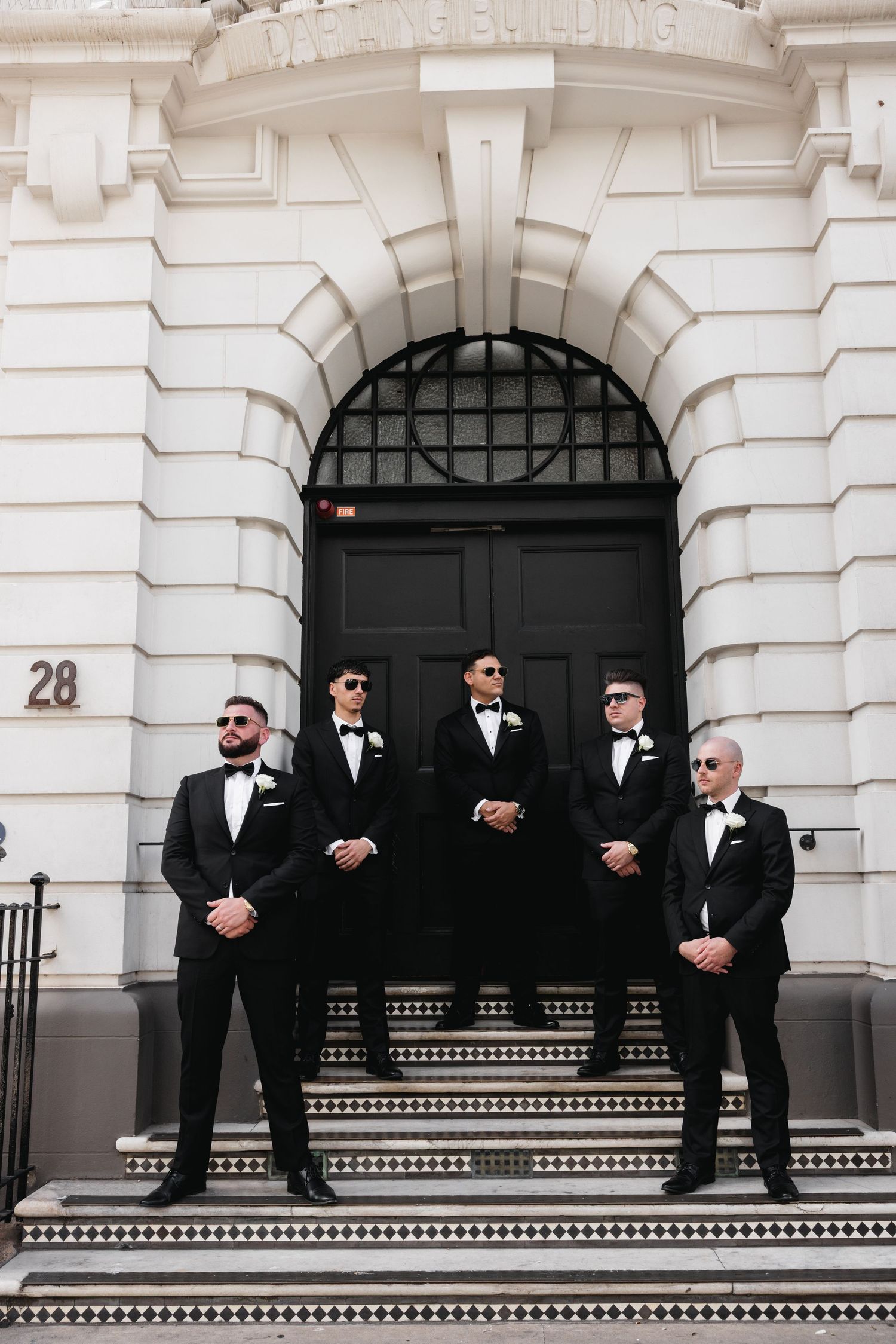 Four groomsmen in black tuxedos with bow ties posing on steps of elegant white building.