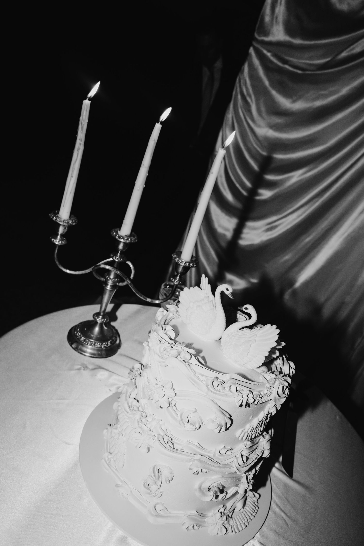 Elegant black and white wedding cake with swan topper beside lit candelabra on draped table.