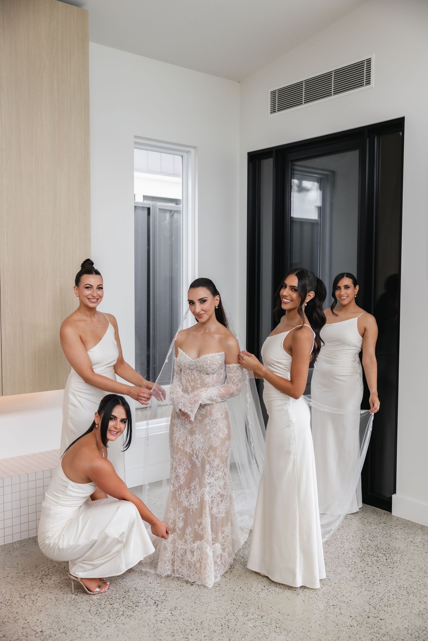 Bride in lace wedding gown surrounded by four bridesmaids in white dresses helping her prepare in a bright modern room.