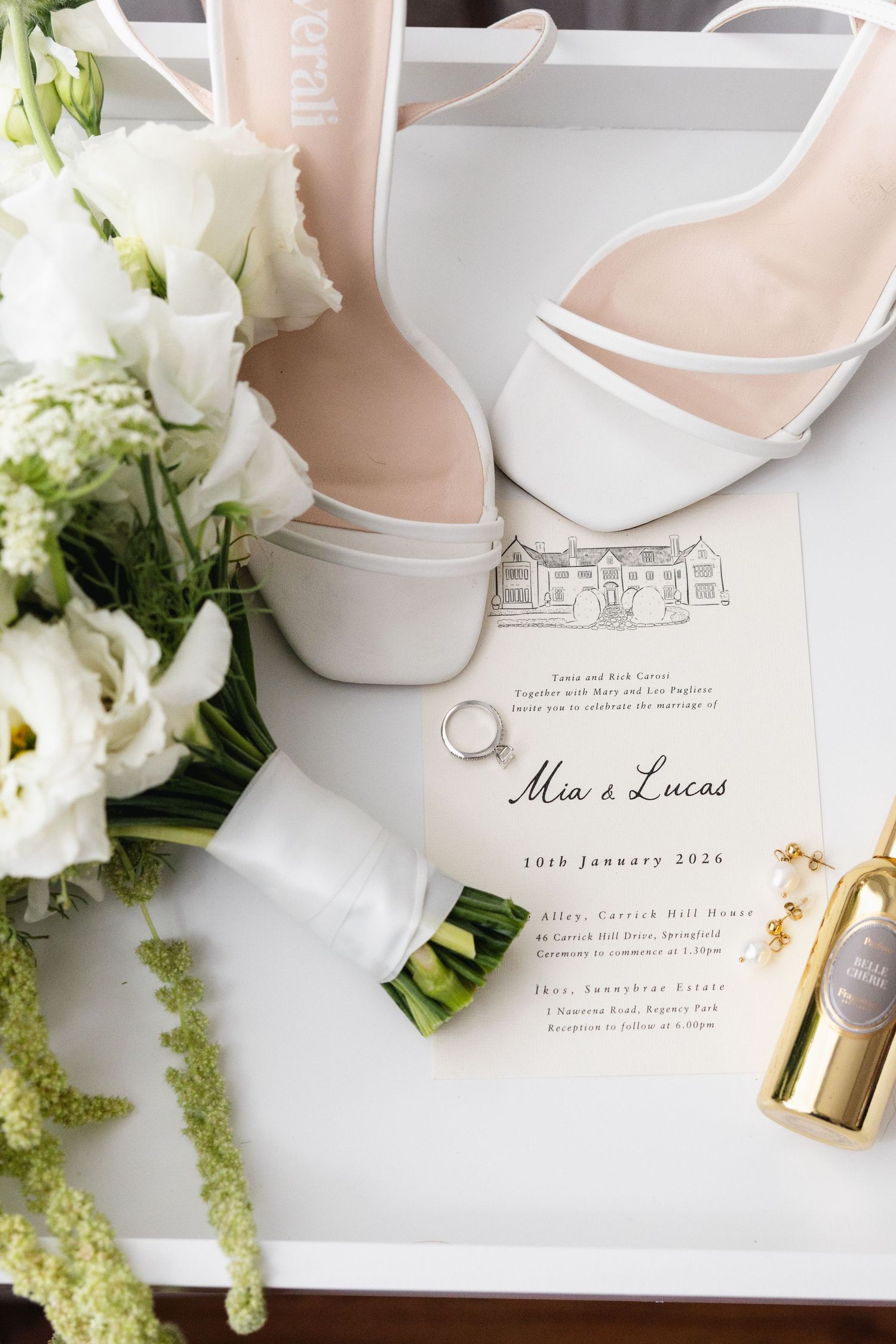 Elegant wedding flat lay featuring white heels, floral bouquet, gold perfume bottle, rings, and Mia & Lucas invitation on white surface.