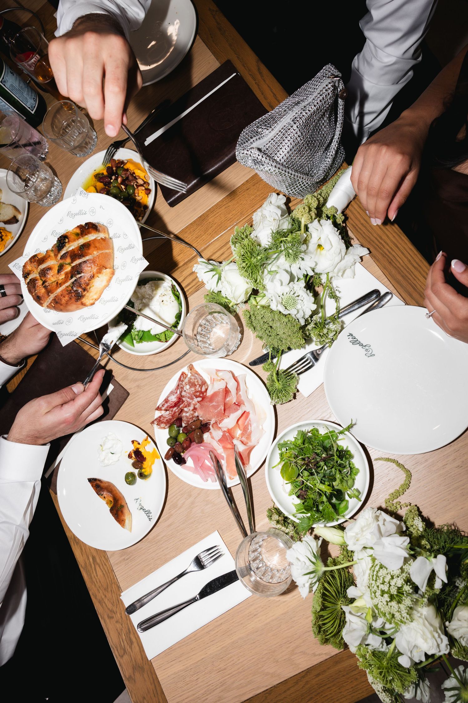 Overhead view of a restaurant table with charcuterie, salad, and bread plates surrounded by white floral arrangements.