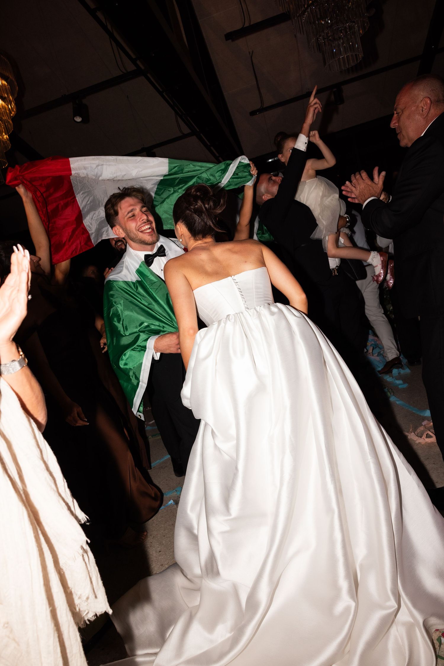 Bride and groom dancing joyfully at wedding reception with Italian flag waved by guests in background.