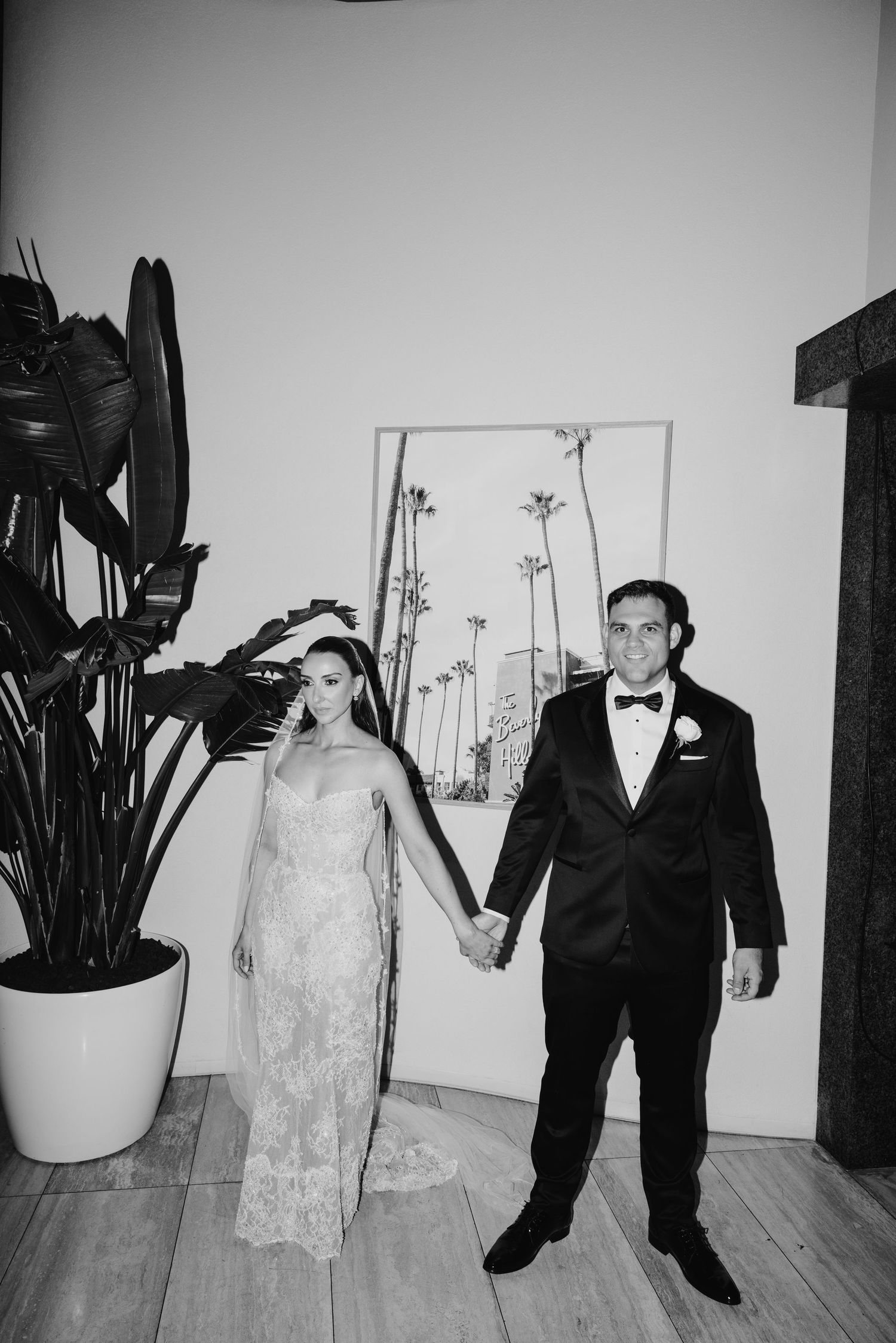 Bride in lace gown and groom in tuxedo holding hands indoors.