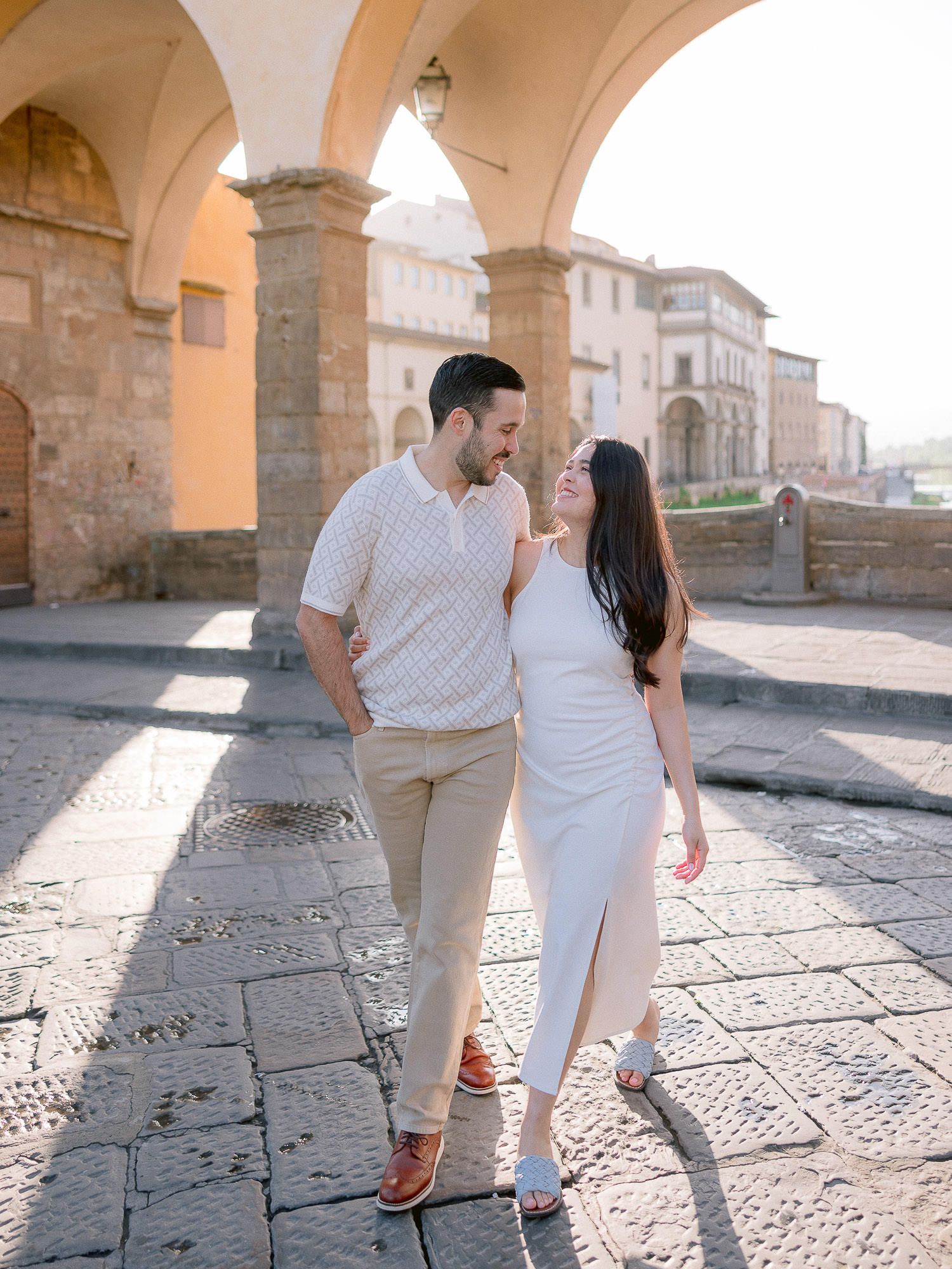 Couple walking hand in hand under stone archways in Florence during golden hour.