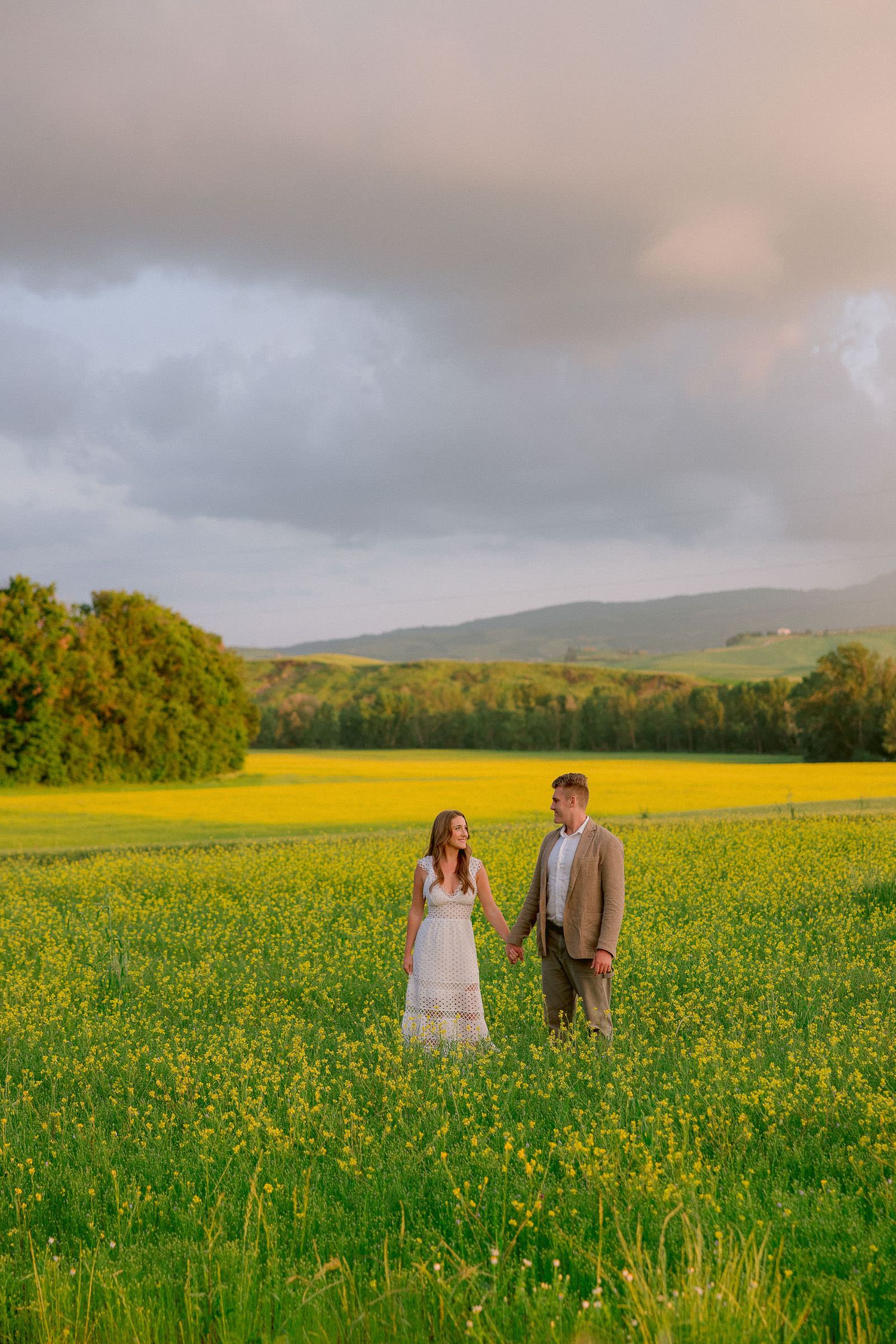 Couple walking through vibrant yellow wildflower field in Tuscan countryside at sunset.