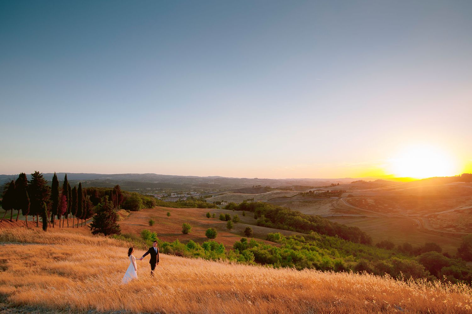 Panoramic sunset view over rolling Tuscan hills with cypress trees in the distance.
