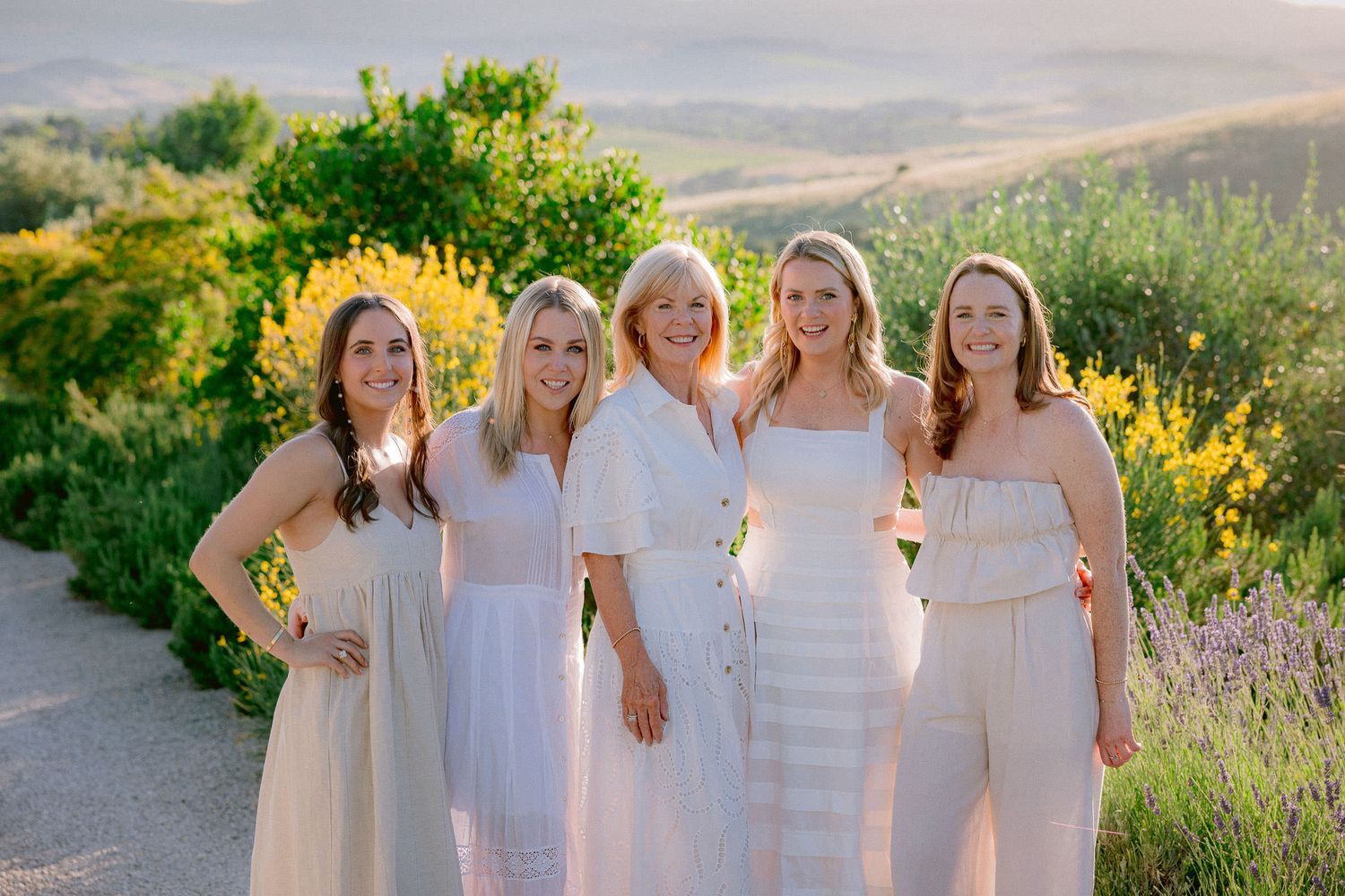 Group wearing white dresses poses together in a sunlit Mediterranean garden.