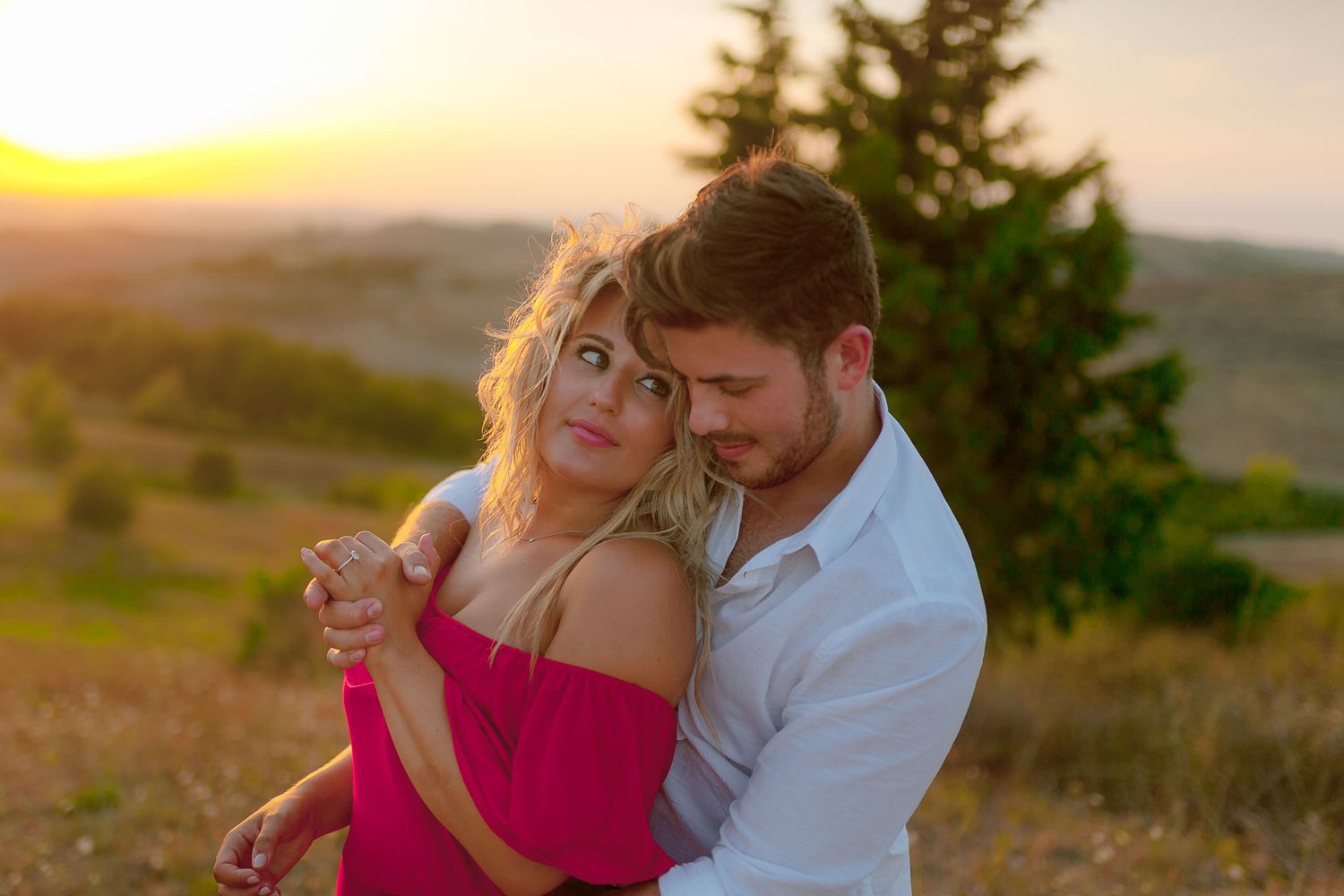 Couple embracing in a romantic sunset moment on a hilltop overlooking countryside.