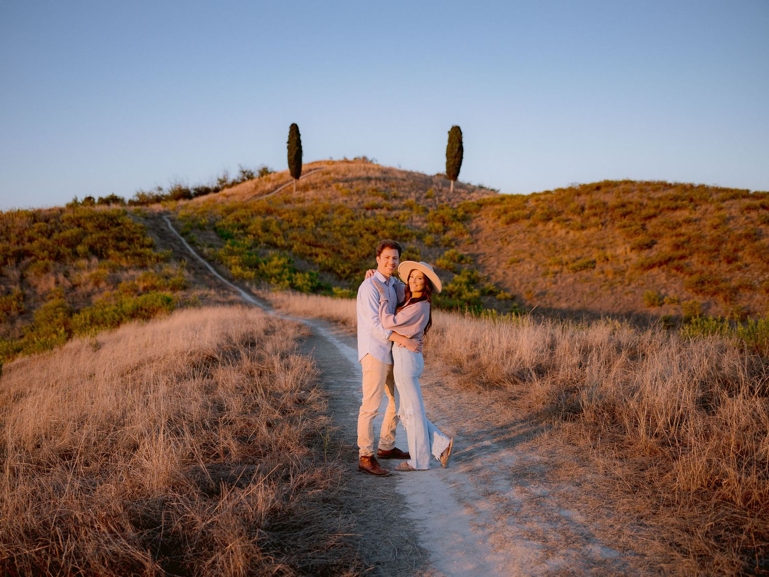 A winding dirt path leads through golden brown hills during sunset in the countryside.