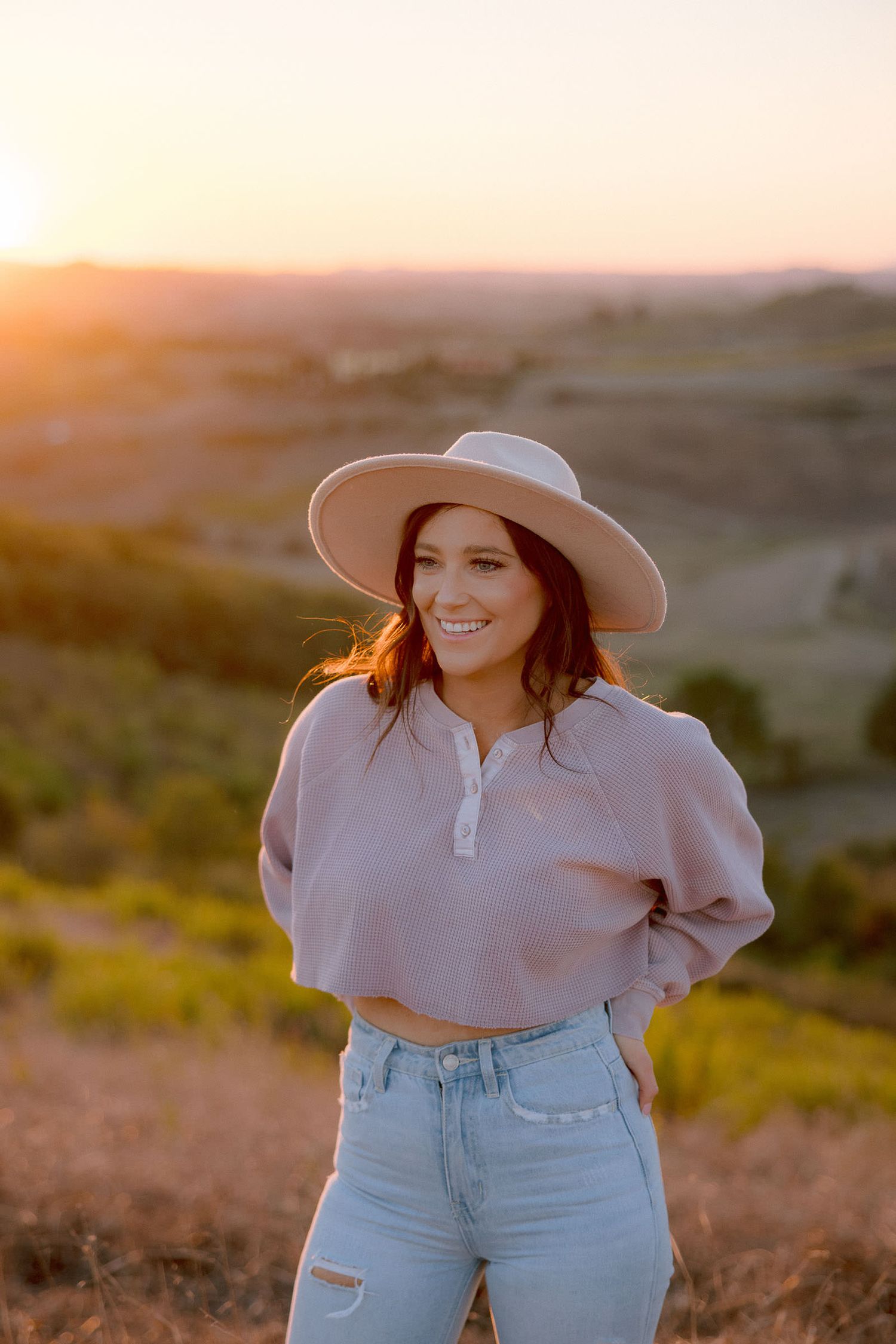 Woman in wide brim hat and casual attire poses against golden sunset landscape.