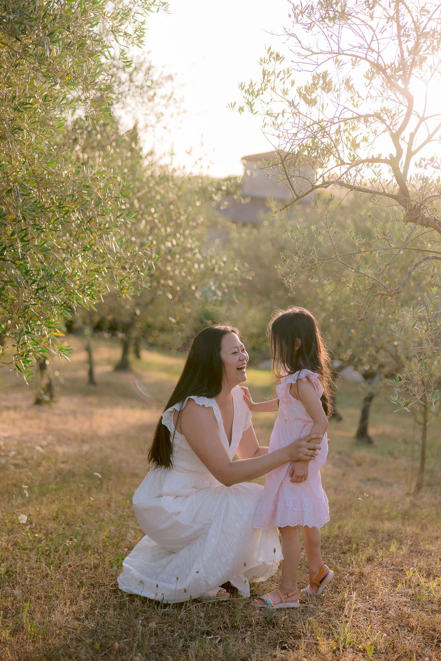 Tender mother-daughter moment captured in white dresses among blooming trees at sunset.